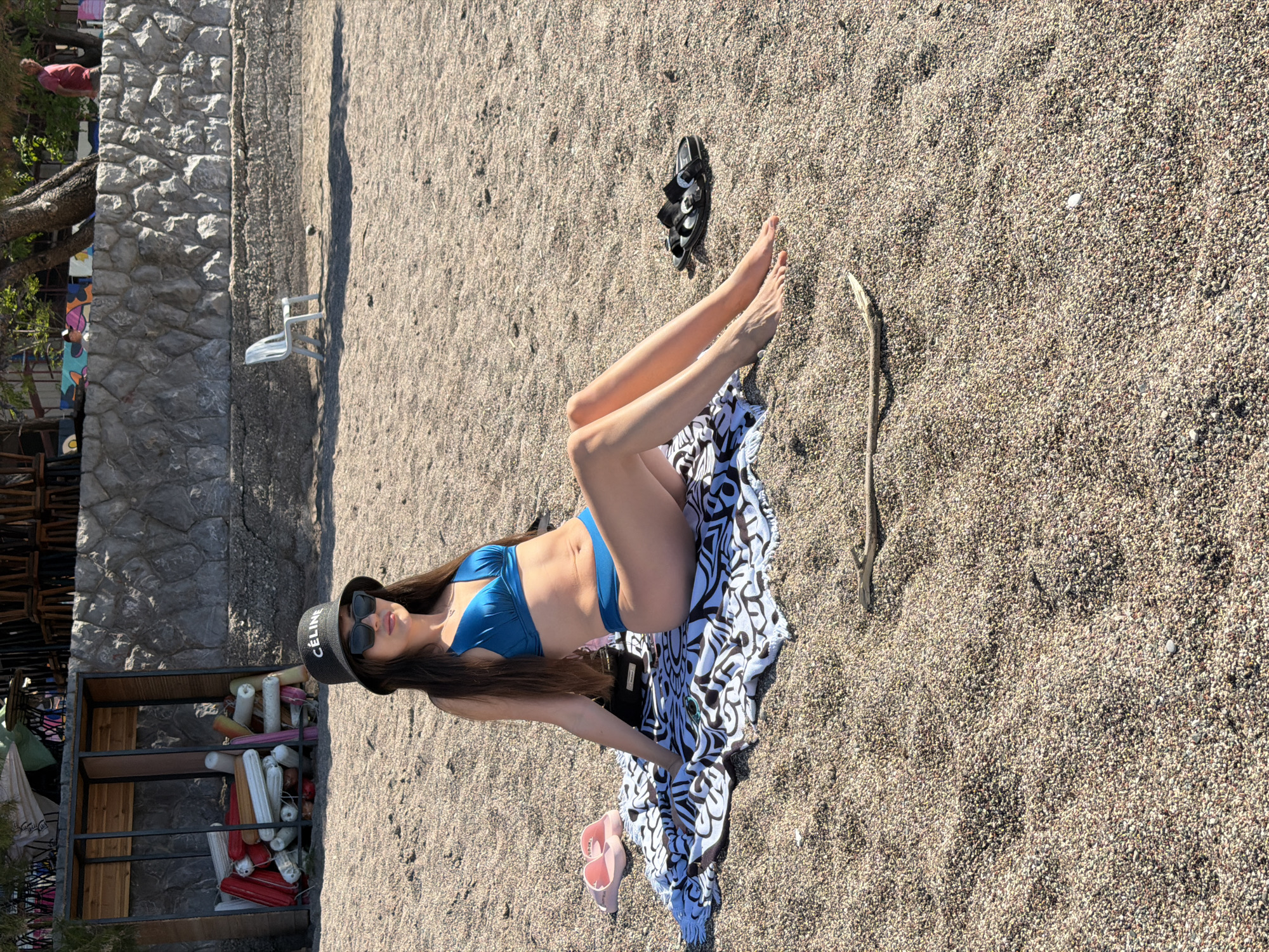 The image displays a young woman relaxing on a sun-drenched, pebbly beach in Rafailovići, Montenegro. She is lying on her back, propped up, with her legs extended and crossed at the ankles. The woman has long, dark hair and is wearing a blue two-piece bikini, black sunglasses, and a black bucket hat that features the brand name "CELINE" in white text.
She is resting on a black and white patterned beach towel spread across the gravelly sand. Beside her towel are pink flip-flops, and a pair of black sandals are visible further up on the beach. A slender piece of driftwood also lies on the sand nearby.
In the background, a stone wall runs along the edge of the beach, with green foliage and trees visible beyond it. To the left, a wooden-framed structure, possibly a storage unit or stall, is filled with numerous colorful, elongated buoys or floats in shades of white, red, pink, yellow, and blue. A white beach chair can be seen in the distance on the sand.
The scene is brightly illuminated by direct sunlight, suggesting late morning or early afternoon, with clear skies and distinct shadows. The overall impression is one of leisure and relaxation on a beach holiday.