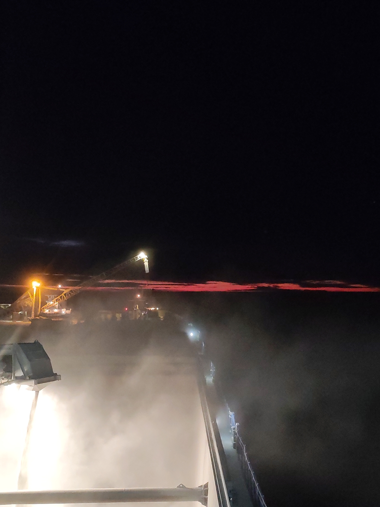 The image shows an industrial scene at night, possibly in or near Shcheleiki, Russia, due to the request in the prompt. There is heavy fog or steam obscuring much of the lower view. In the distance, an elevated platform or crane is visible with lights on. Reddish streaks can be seen on the horizon, possibly indicating a sunset or the glow from a nearby industrial activity. In the foreground, there is a structure, likely a building or a platform, which appears to be the vantage point of the image. There are no visible people. The presence of industrial equipment suggests this may be a factory, power plant, or other similar facility.