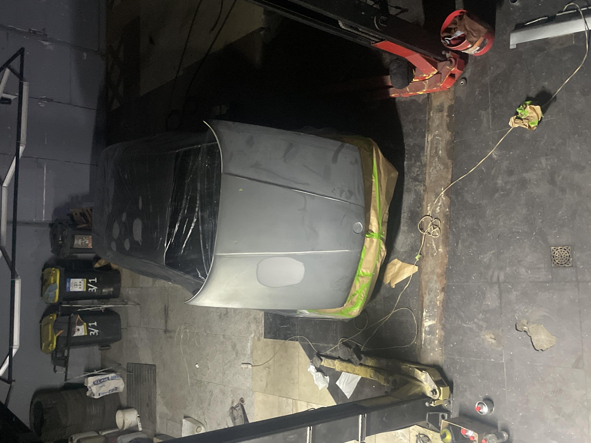 In an indoor garage, a car hood is raised and covered in plastic wrap. The hood appears to be undergoing some form of automotive repair or preparation for painting, with a lighter colored patch, possibly primer or filler, visible. The car is positioned on a car lift, and paper and plastic sheeting are used to mask off the surrounding areas. Various tools and materials, including spray cans, rags, and trash bins, are scattered around the workspace. The floor is concrete and shows signs of wear and tear. A red hydraulic jack is visible to the right of the vehicle. The lighting in the garage is dim, with overhead fluorescent lights providing illumination. The overall impression is of an active auto repair or restoration area.