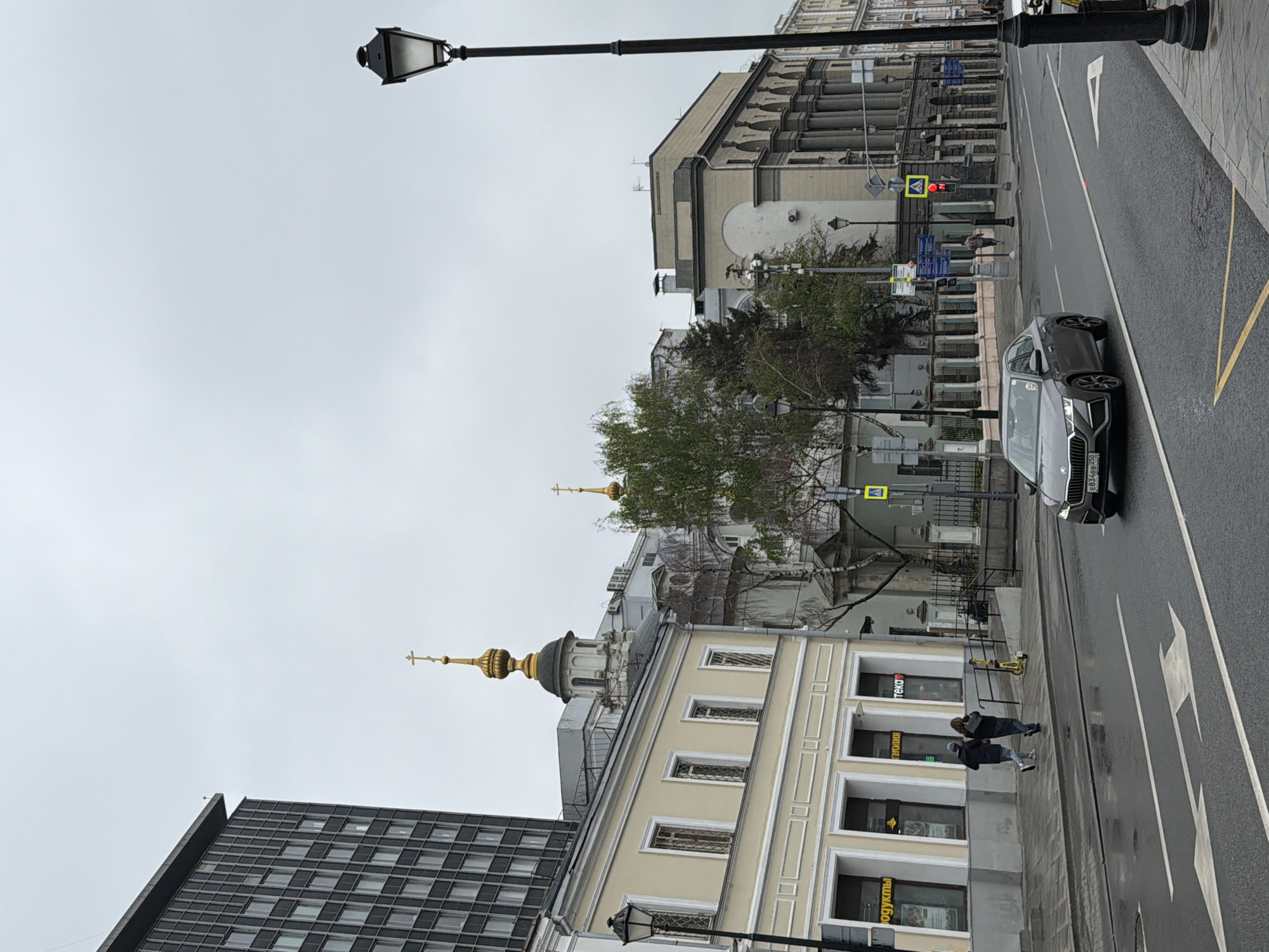 The image depicts a street scene in Moscow, Russia on a cloudy day. A dark grey Skoda car is parked on the right side of the road, facing forward. Two pedestrians are walking on the sidewalk next to a light yellow building, which has a golden dome and cross visible above it. The building appears to be a church or religious building. A tall, dark grey modern building is visible to the left of the light yellow building. In the background, there are older buildings with columns and architectural details, as well as a tree. A black lamppost stands tall on the left side of the road. The street is wet, suggesting recent rain. A pedestrian crosswalk sign is visible, with the light indicating a red light, suggesting pedestrians should not cross.