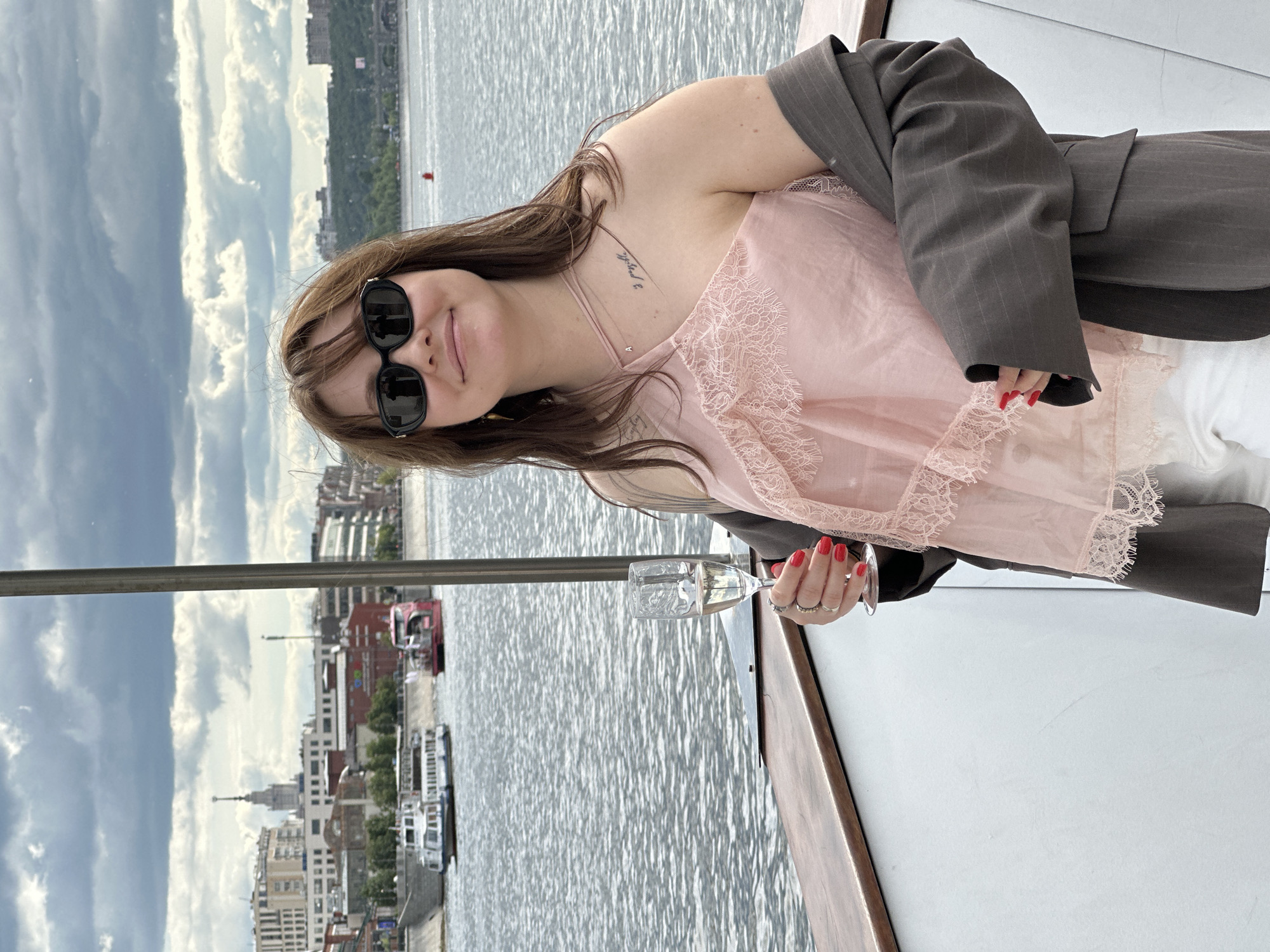 A woman wearing sunglasses, a pink top with lace trim, and a gray blazer stands on a boat holding a glass of champagne. The backdrop features the skyline of Moscow, Russia, including buildings and water. The sky is cloudy.