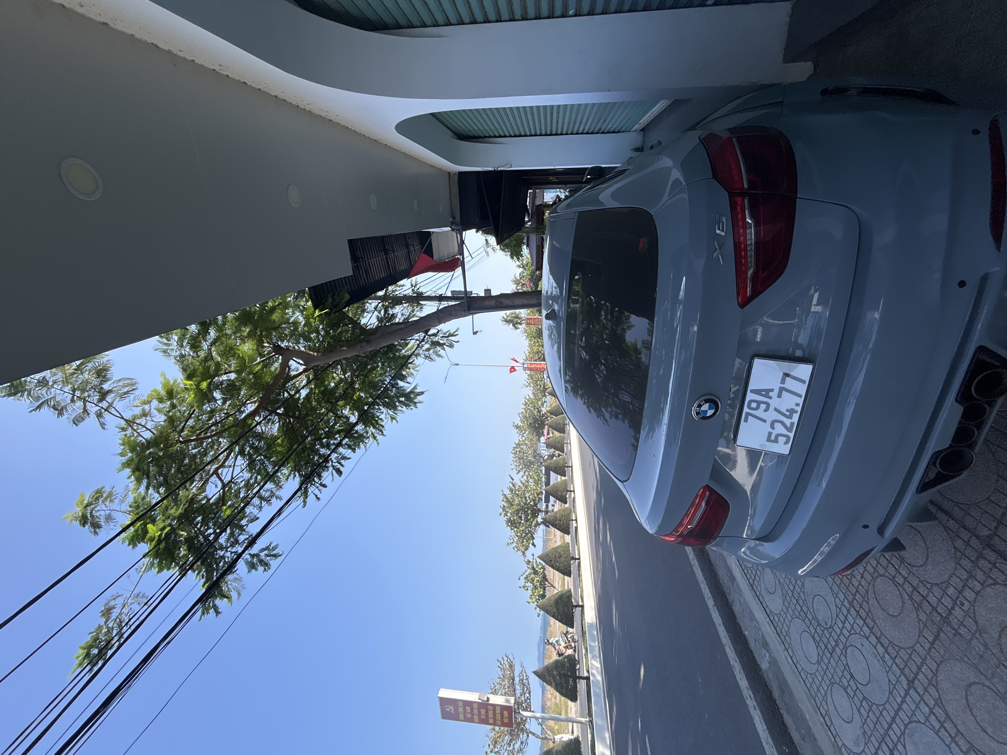 The image captures a sunny daytime scene featuring a grey BMW SUV, likely an X6, positioned on a paved road next to a modern building. The perspective is slightly elevated, looking towards the rear of the vehicle and down the road.
The car, light grey or silver in color, occupies the right side of the frame, showcasing its red taillights and a dual exhaust system. A white Vietnamese license plate, "79A 524.77", is prominently displayed on the rear. The BMW emblem is visible above the license plate.
To the left, a building features a light grey, possibly concrete, overhang with several recessed circular lights embedded in its underside. A green slatted garage door or awning is visible behind the car, suggesting a covered area. Beyond the building, a large tree with lush green foliage extends across the upper left of the image. Numerous black electrical or utility cables are strung across the clear blue sky, intersecting the tree's branches.
Along the right side of the road, a row of uniformly trimmed, conical-shaped bushes lines the pavement. In the distance, a body of water, likely the sea, stretches to the horizon, with a coastline and scattered greenery. Small figures, possibly individuals on motorcycles, are visible along the distant road. A sign post with red and white text is also present further down the road, and a red flag flies from a pole in the mid-ground.
The weather is bright and clear, indicative of a sunny day with a deep blue sky. The sidewalk features patterned grey tiles.
Visible text includes the car's license plate, "79A 524.77". Text on the distant sign post, as identified by OCR, reads "BẢNG CỘNG SẢN", "KIỆT NAM", "TẾ CHỨC", "NƠI TRÁNG LỢI", "CỦA CÁCH MANG", and "BET NAM". These phrases suggest a political or nationalistic sign, consistent with public signage in Vietnam. The location is confirmed as Bắc Nha Trang, Vietnam.