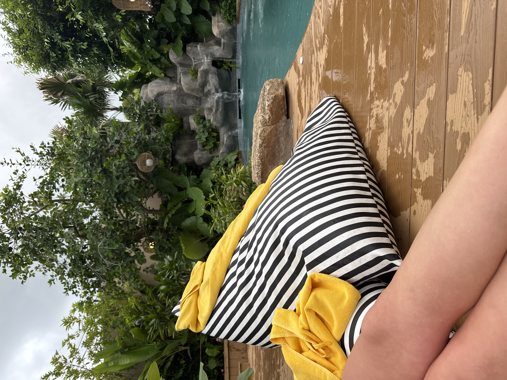 In Chalong, Thailand, a person's legs are visible in the foreground, resting on a wet wooden deck. Beside them is a large, black and white striped beanbag chair with yellow towels draped over it. The beanbag and deck are next to a bright blue swimming pool, which is partially obscured by the beanbag. In the background, lush green tropical foliage surrounds the pool, and a small waterfall cascades down a rocky embankment. The sky above is overcast.