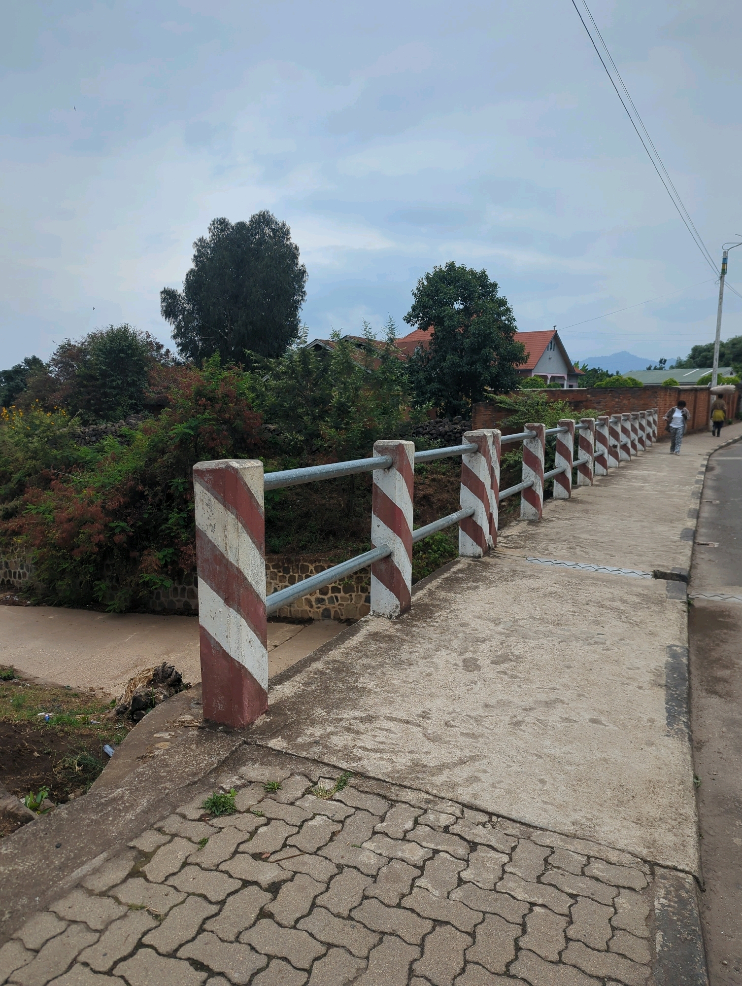 The image shows a view of a bridge railing and sidewalk in Musanze District, Rwanda. The railing is made of metal bars supported by concrete posts painted with red and white diagonal stripes. The sidewalk is paved with interlocking concrete blocks in the foreground and smooth concrete further back. To the left of the bridge, there is a dry riverbed or ditch with some vegetation and debris. Beyond the railing, there is a dense green hillside with trees and some buildings, including houses with red-tiled roofs. In the background, mountains are visible under a cloudy sky. Two people are walking on the sidewalk in the distance, one in a white top and patterned pants, and another with a yellow backpack.