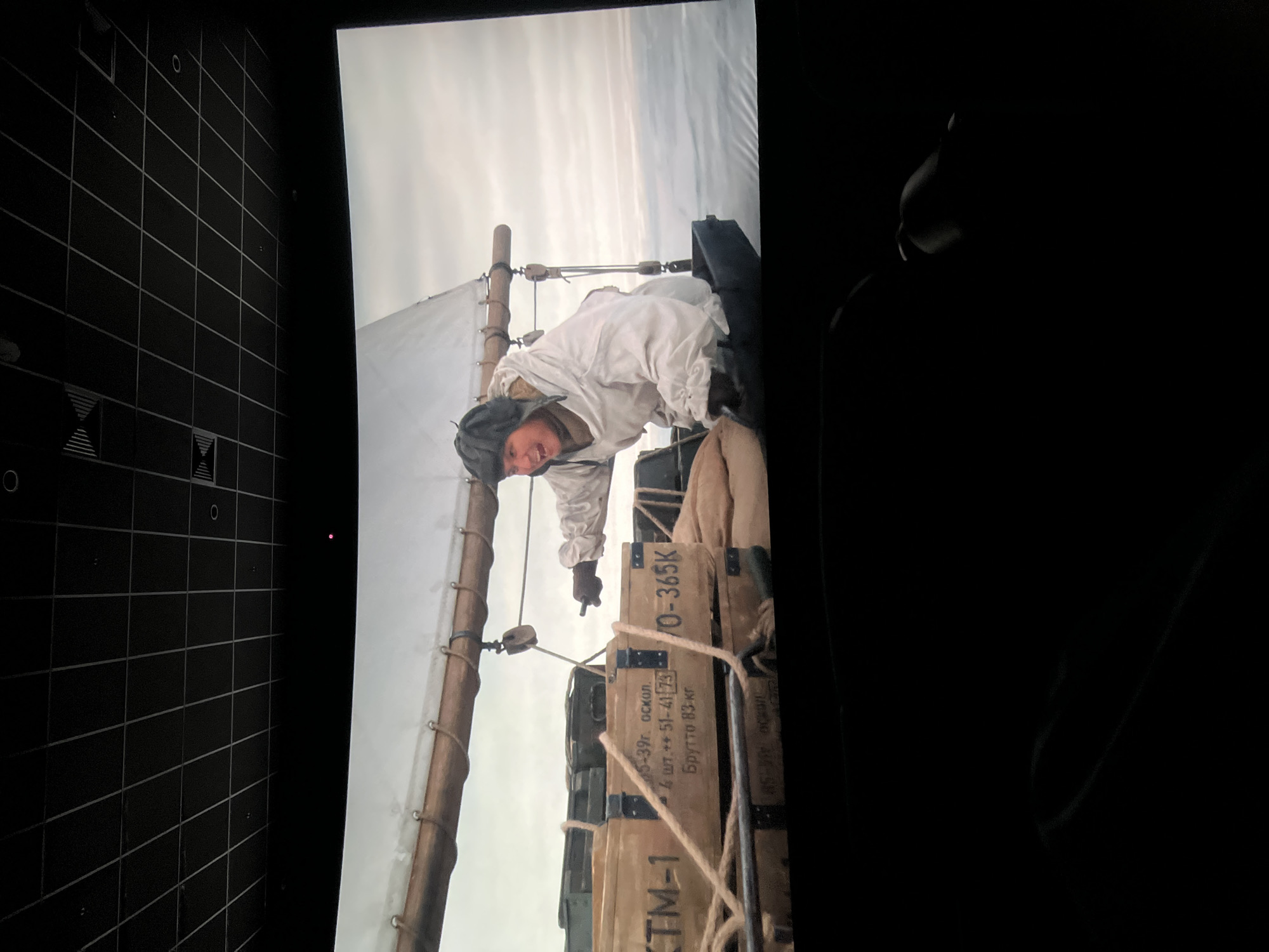 The image shows a scene from the movie "The Best in Hell." A man in a white winter uniform is climbing on top of wooden crates labeled "TM-1", "5-39r. ockon," "4 uT ++ 51-4173," "Brytto 83-Kr," and "O-365K." The location in the city of Krasnoyarsk, Russia is not discernible from the image alone.