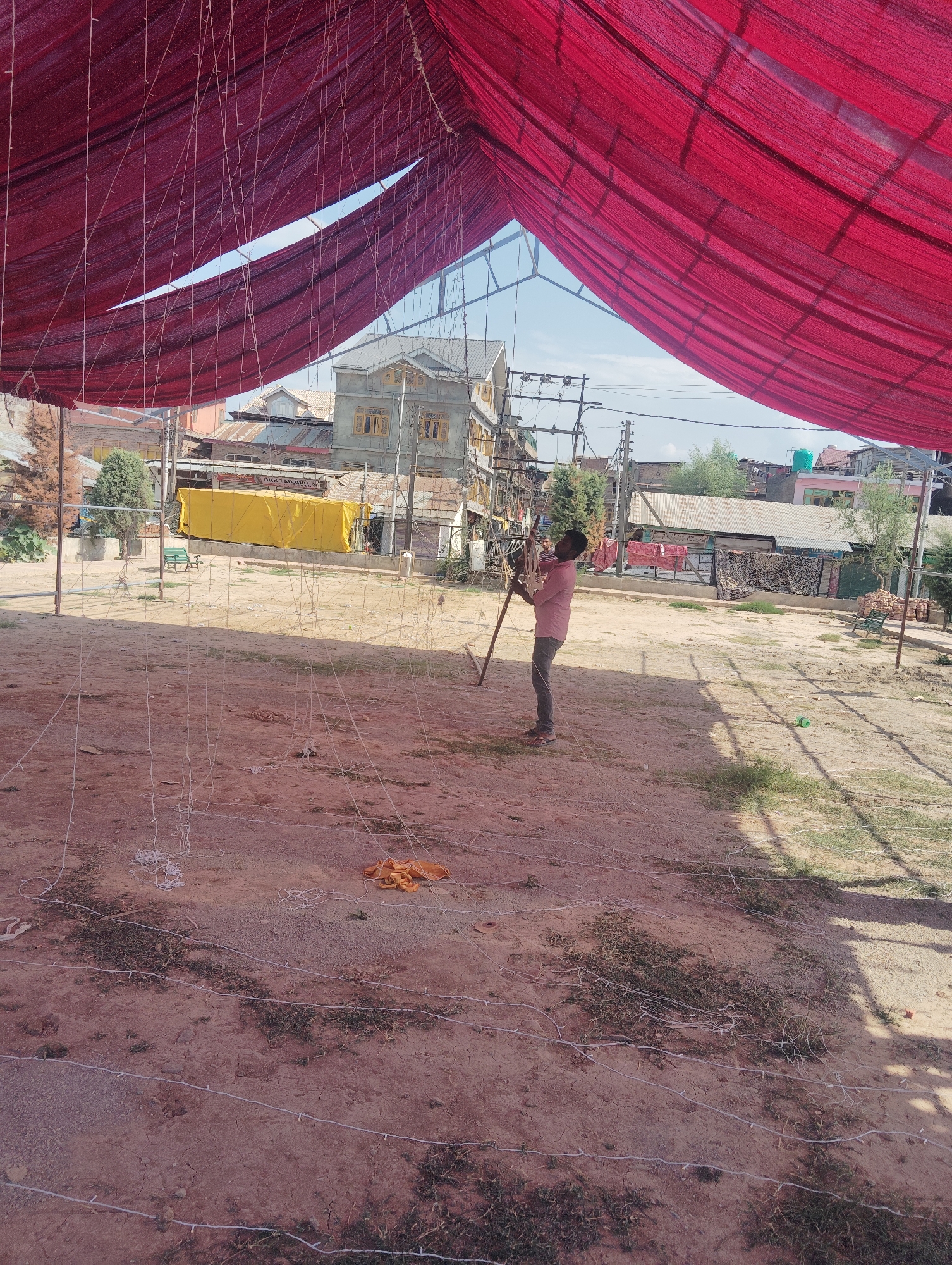 A man in a pink shirt and gray pants is seen working with a long wooden pole in an outdoor, dusty area in Srinagar, India. He appears to be engaged in setting up or dismantling a large red tent, the fabric of which hangs overhead, creating a canopy. The ground is covered with dirt and scattered white strings, possibly part of a decorative light setup. In the background, buildings, trees, and power lines suggest an urban or semi-urban environment. The overall scene depicts an ongoing task related to event setup or maintenance under a clear sky.