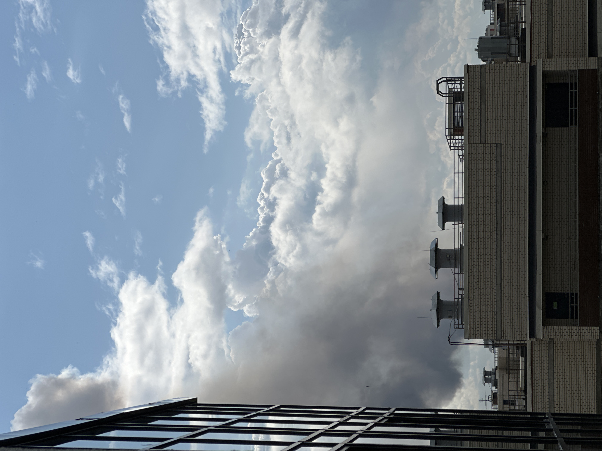 The image, captured from a high vantage point, likely from a window or balcony in the city of Dnp Ryzhiki, Russia, depicts a rooftop scene dominated by a large, dark plume of smoke billowing into a partly cloudy sky. The smoke originates from behind several rooftop structures, obscuring a portion of the view. In the foreground, the edge of a modern building with large, dark-framed windows is visible on the left side of the frame, reflecting the sky and the smoke.
The rooftops in the mid-ground are characterized by a light-colored brick construction. Several ventilation units, appearing as large cylindrical structures with caps, are prominent features on these rooftops. Some of these units are connected by pipes and wiring. Metal railings and structural elements are also visible on the rooftops, suggesting industrial or functional spaces. The background is filled with a dramatic sky featuring a mix of bright white, fluffy cumulus clouds and a lighter blue sky. The smoke plume is a significant element, creating a stark contrast with the bright clouds and blue sky. There are no visible people in the image. The overall impression is one of an urban industrial environment under a dynamic sky, with the smoke suggesting an ongoing event.