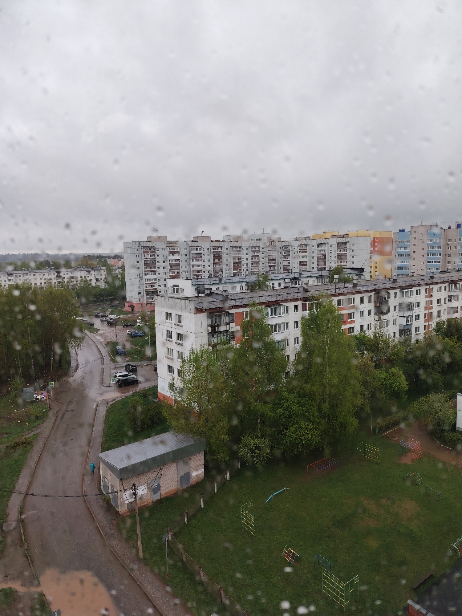 A rainy, overcast day in Chaikovsky, Russia, as viewed from a high vantage point, likely a window, evident from the water droplets on the glass. The scene depicts a typical residential area with multiple Soviet-era apartment buildings of varying heights and colors: predominantly white and grey, some with orange or blue accents. A network of wet roads and sidewalks suggests recent rainfall.
To the left, a small brick building with a grey roof sits adjacent to a path where a person in a blue coat is walking. Beyond the building is a street with parked cars. A children's playground with various climbing structures and apparatus is visible in the lower right corner, surrounded by green grass. Green trees are interspersed throughout the landscape. The sky is uniformly grey, indicating heavy cloud cover. The overall scene conveys a sense of urban life in a post-Soviet landscape on a wet day.