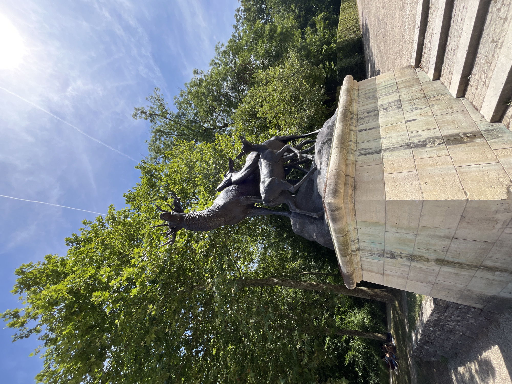 The image shows a bronze sculpture of deer on a stone pedestal, located in Parc de Sceaux, in the city of Antony, France. The sculpture depicts a large stag with its head raised and mouth open, appearing to bellow, with two smaller deer standing beside it. The deer are positioned on a rocky outcrop that forms part of the sculptural composition. The pedestal is made of large, light-colored stone blocks, some of which show staining from water runoff. Steps lead up to the right side of the pedestal. The background is dominated by lush green trees, with a bright blue sky above featuring some wispy white clouds and contrails from an aircraft. In the lower left corner of the image, there is a stone retaining wall, and beyond that, a grassy area where several people are sitting and lying down. The overall scene suggests a sunny day in a park.