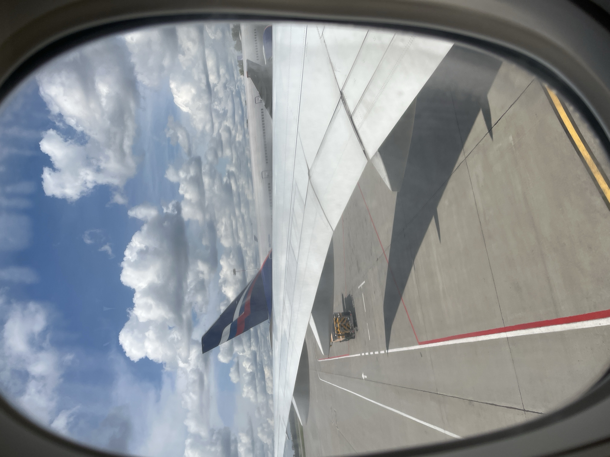 An Aeroflot airplane is parked on the tarmac at Rusanovo Airport in Rusanovo, Russia, under a partly cloudy sky. The view is from inside another aircraft, looking out the window at the wing of the parked plane. The wing is white and extends across the foreground of the image, casting a shadow on the tarmac. In the background, the tail fin of the Aeroflot plane is visible, featuring the airline's distinctive red and white logo. A yellow baggage tug is positioned on the tarmac near the wing. The tarmac is marked with white and red lines, indicating taxiways and parking areas. The scene suggests an airport environment with ongoing operations.