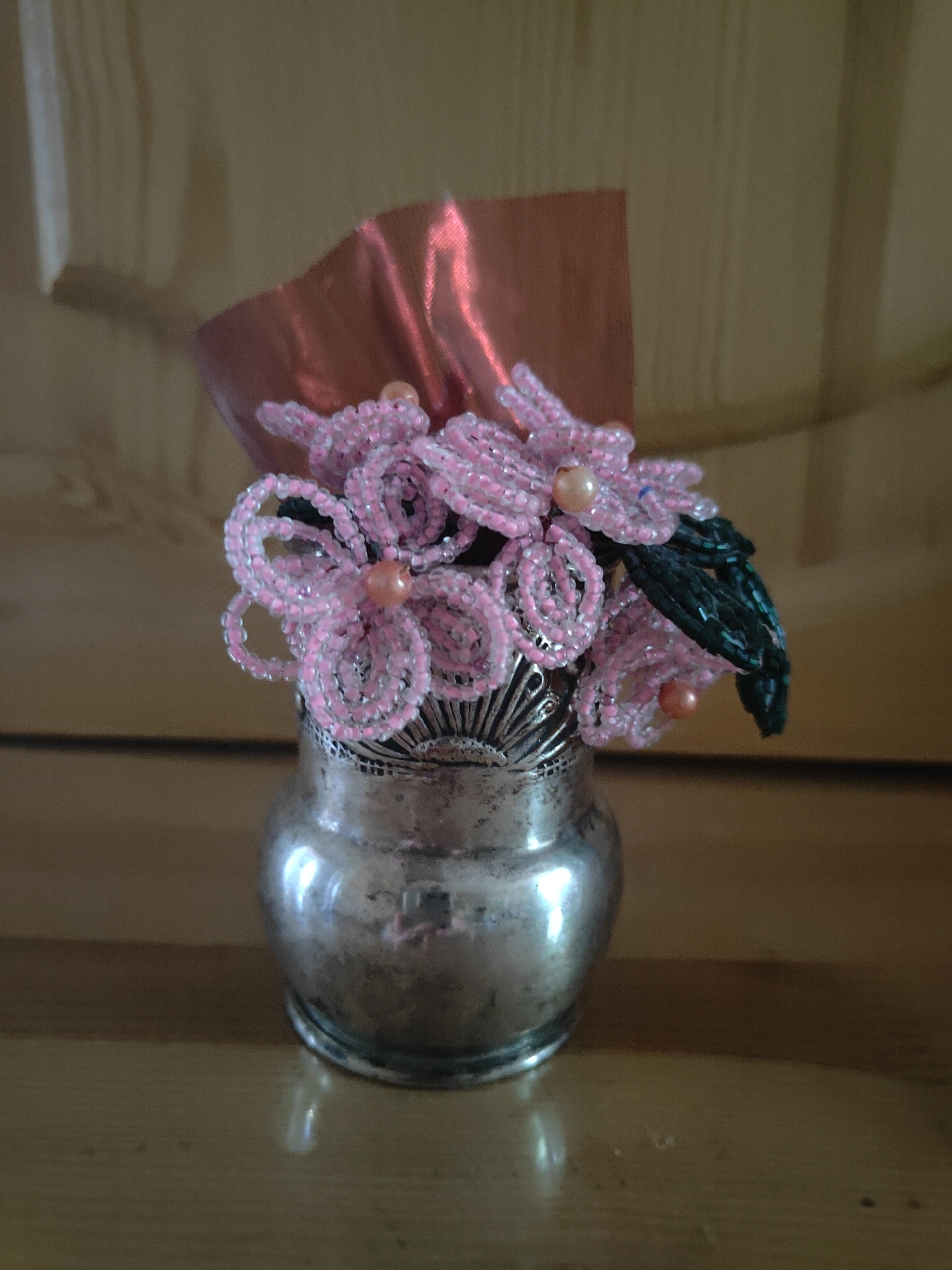 A still life image captures a small, ornate silver vase filled with delicate pink beaded flowers and greenery. A piece of shimmering pink fabric is tucked behind the flowers. The vase is metallic and appears slightly tarnished, with intricate detailing around its rim. The flowers are intricately crafted from small pink beads, with a few larger, peach-colored beads serving as the flower centers. Dark green beads form the leaves. The vase sits on a polished wooden surface, and the background is a muted, textured wall. The lighting is soft, casting gentle shadows. There are no people present in the image. The location is identified as Unknown, Unknown.
