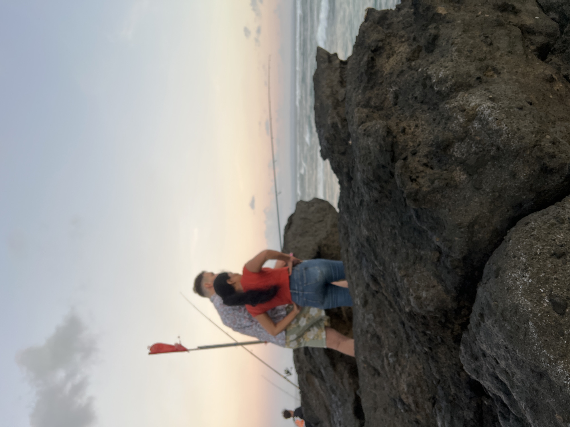 At dusk on the rocky shores of Mengwi, Indonesia, a couple stands on large, dark volcanic rocks near the water's edge. The man, wearing a patterned short-sleeved shirt and patterned shorts, stands behind a woman dressed in a red sleeveless top and blue jeans. She has her hands on her hips, looking out at the ocean. Several fishing rods are visible, some planted in the rocks. A bright red flag on a pole is positioned to the left of the couple. The sky above is a soft gradient of blue and orange, with a few wispy clouds. The sea is active with waves crashing against the shore. In the background, a person with dark hair is also visible, adding to the sense of activity at this coastal location.