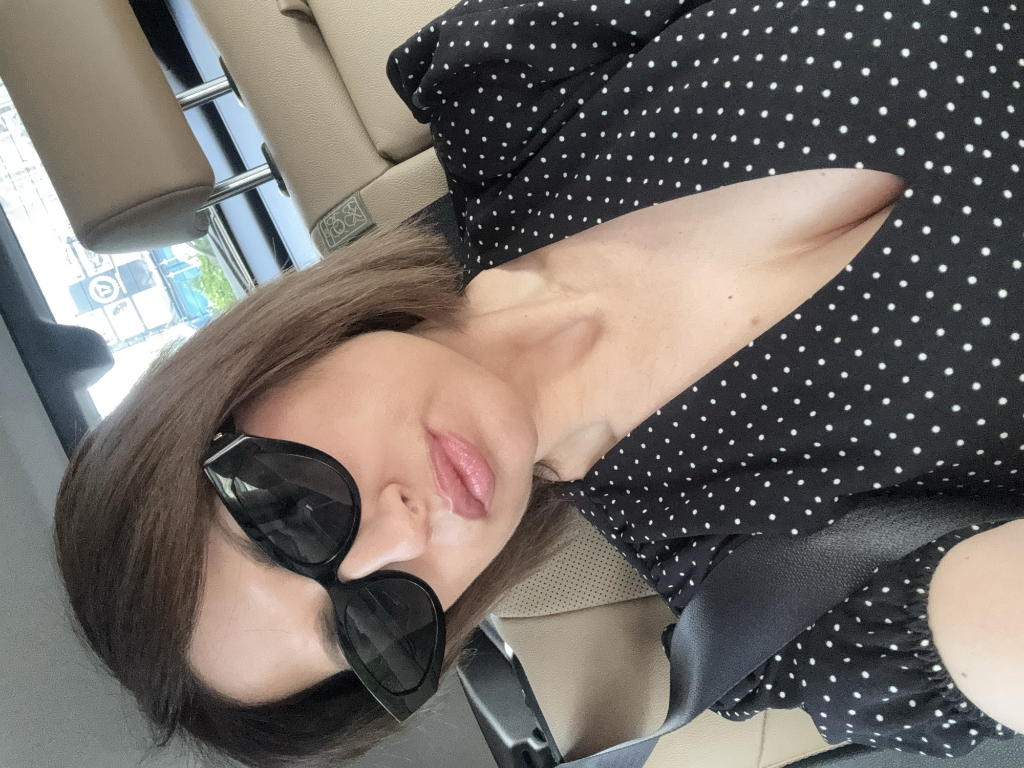 A woman is depicted inside a vehicle, likely a car, positioned at a tilted angle. She has medium-length, light brown hair and is wearing large, black, cat-eye style sunglasses. Her lips are slightly parted, revealing a light pink lip color. She is dressed in a black top or dress adorned with small white polka dots, and a black seatbelt is visible across her chest and shoulder. The interior of the vehicle features beige upholstered seats and headrests. Through the window, parts of an urban environment with buildings and what appears to be a street are visible, suggesting it is daytime. On the headrest upholstery, a small, light-colored emblem or logo can be seen, which appears to be a stylized design, possibly containing letters or numbers like "18x" or "XO" within a rectangular outline. The woman is looking towards the camera, possibly taking a selfie. The overall setting is Sofia, Bulgaria.