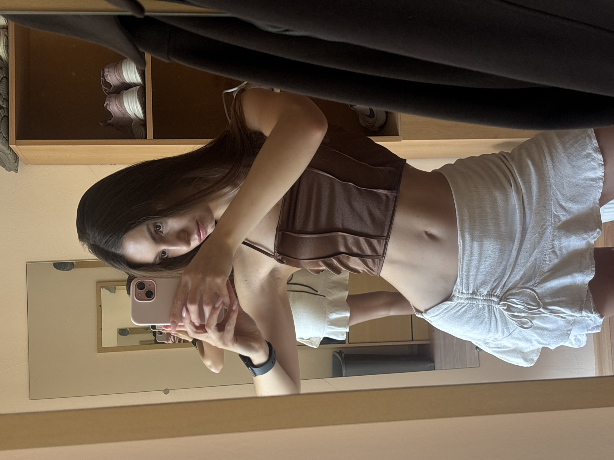 A young woman with long dark hair takes a mirror selfie. She is wearing a brown corset-style top and a white linen skirt with a drawstring. The woman is leaning forward, holding a pink smartphone to capture the image. In the background, a wooden shelf unit is visible, containing a pair of light pink and white sneakers. A full-length mirror reflects the woman and the immediate surroundings. The overall setting appears to be a domestic space, possibly a bedroom or dressing area. The lighting is soft, suggesting it could be daytime. The woman's expression is neutral. There is no visible text in the image. The location context is Varna, Bulgaria.