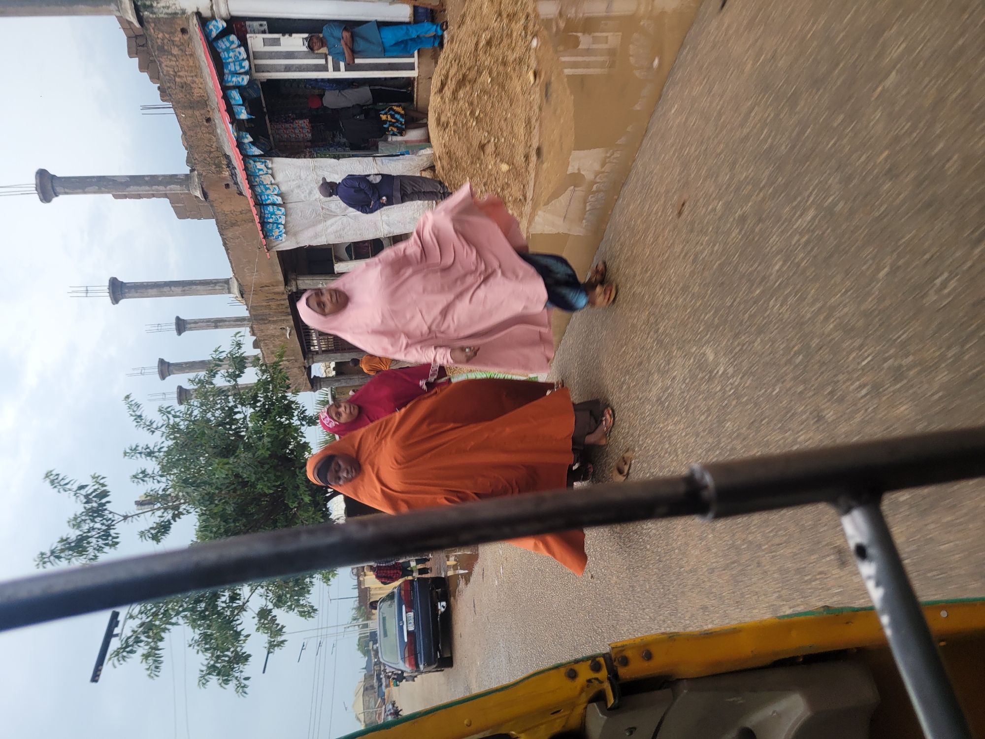 The image, taken from a moving vehicle, captures a street scene in Jos, Nigeria, characterized by an overcast sky. In the foreground, a portion of a yellow and green motorized tricycle is visible, along with its black metal frame, indicating a passenger's perspective. Three women are the central figures, walking on the asphalt road. The woman closest to the camera, on the right, wears a flowing pink hijab and matching dress, her gaze directed forward. To her left, another woman is dressed in an orange hijab and dress, her head bowed. A third woman, partially obscured, is in a dark red hijab and dress.
Behind the women, a dark blue sedan is parked on the side of the road, its license plate visible. The road itself appears wet, with large puddles suggesting recent rain. To the right of the road, a construction site is evident, with a partially built structure featuring concrete pillars and brickwork. In front of this structure is a small shop with goods displayed outside. Several other individuals are present, including men in blue and dark clothing standing near the shop, and others further in the background. A pile of sand is also present on the construction site, near the flooded area. Trees with green foliage are scattered throughout the scene, and streetlights are visible on poles in the distance.