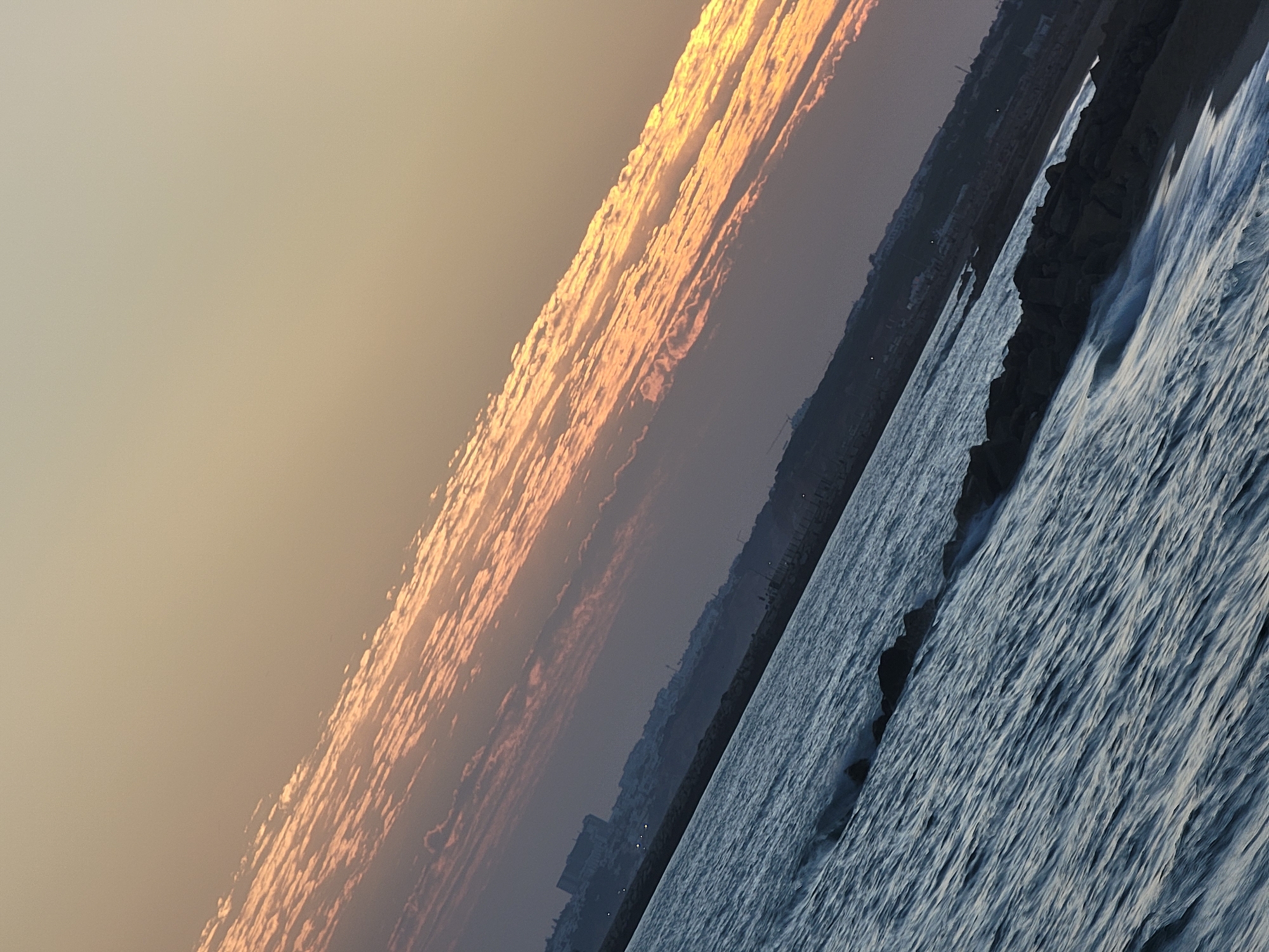 The image depicts a diagonal view of the ocean and the coastline of Quarteira, Portugal, at sunset. The sky is a gradient of pale orange and yellow, with long, wispy clouds illuminated by the setting sun, creating a streaked effect. Below the sky, the horizon is a dark silhouette of the city, with buildings and structures visible. The ocean occupies the foreground, with deep blue waves creating a textured surface. A line of dark rocks, acting as a breakwater or part of the coastline, is visible along the shore, with small waves crashing against them. The lighting suggests it is late afternoon or early evening. There are no people visible in the image.