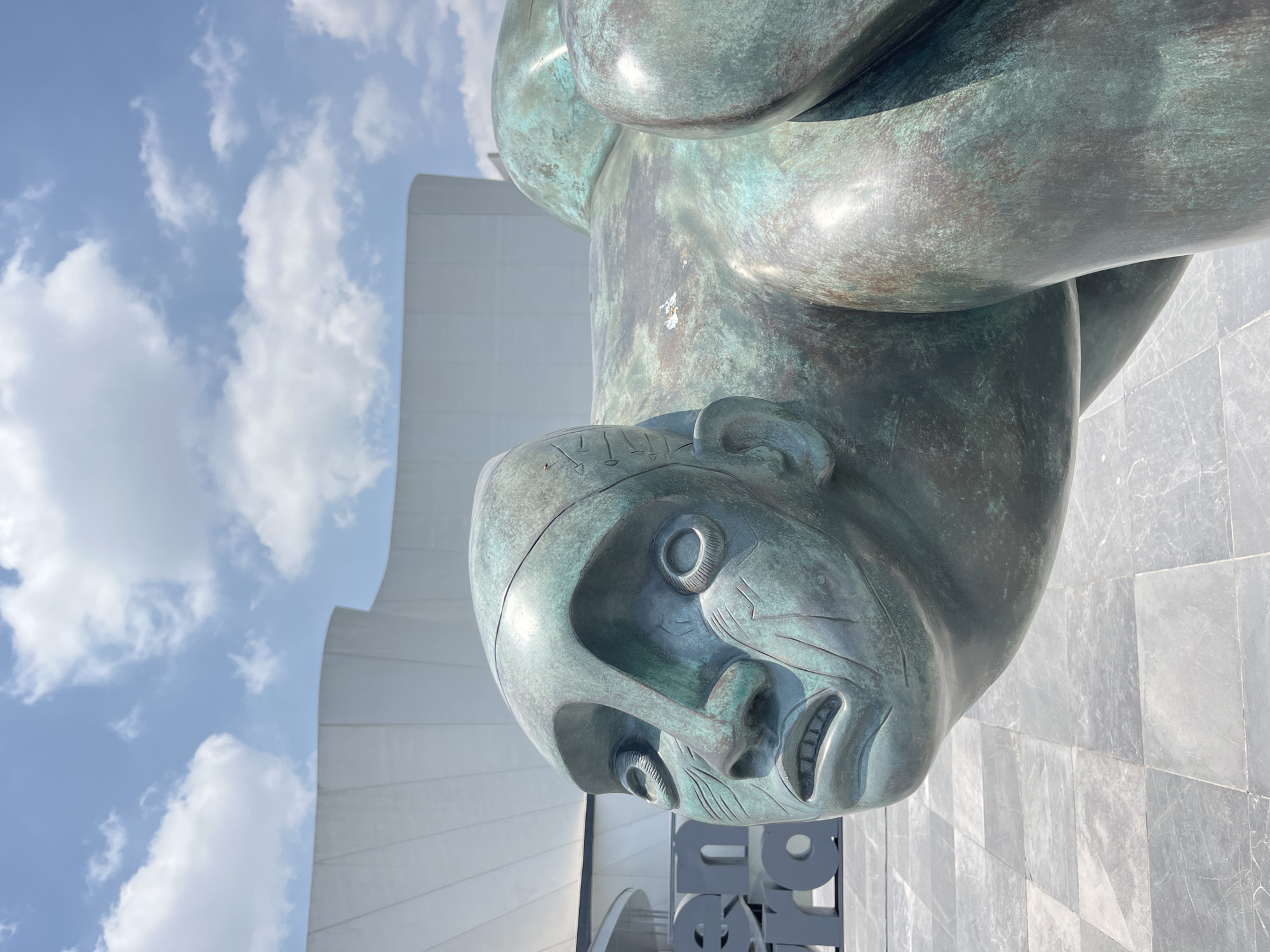 A close-up, low-angle shot showcases a weathered bronze sculpture of a stylized, human figure. The sculpture's face is highly detailed, with prominent eyes, nose, and mouth, suggesting a distinct expression. The body appears bulky and stylized. The statue is located outdoors, with a modern, white building featuring curved architectural elements visible in the background. The sky is partly cloudy, with a mix of blue sky and scattered white clouds. A dark-colored, blocky sign with indiscernible letters is partially visible at the base of the statue. The location is San Bernardino Tlaxcalancingo, Mexico.