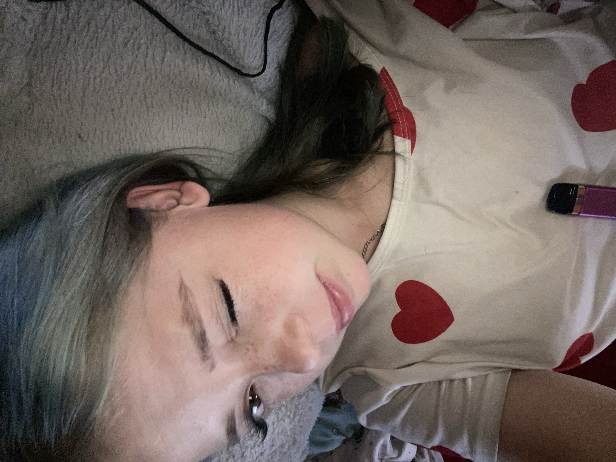 A young female with light skin and dark hair, styled with blue highlights, is lying down on a textured, gray blanket. She is winking with her right eye, her left eye is open, and her lips are slightly parted. She is wearing a light-colored t-shirt with red heart designs. A small, purple electronic device is visible on the lower portion of her t-shirt. The background consists of the gray blanket and a hint of what appears to be bedding or clothing in shades of blue. The image is taken from a close-up, high-angle perspective, with the subject's face being the primary focus. There is no visible indication of the city of Nova Praha, Ukraine within the image.