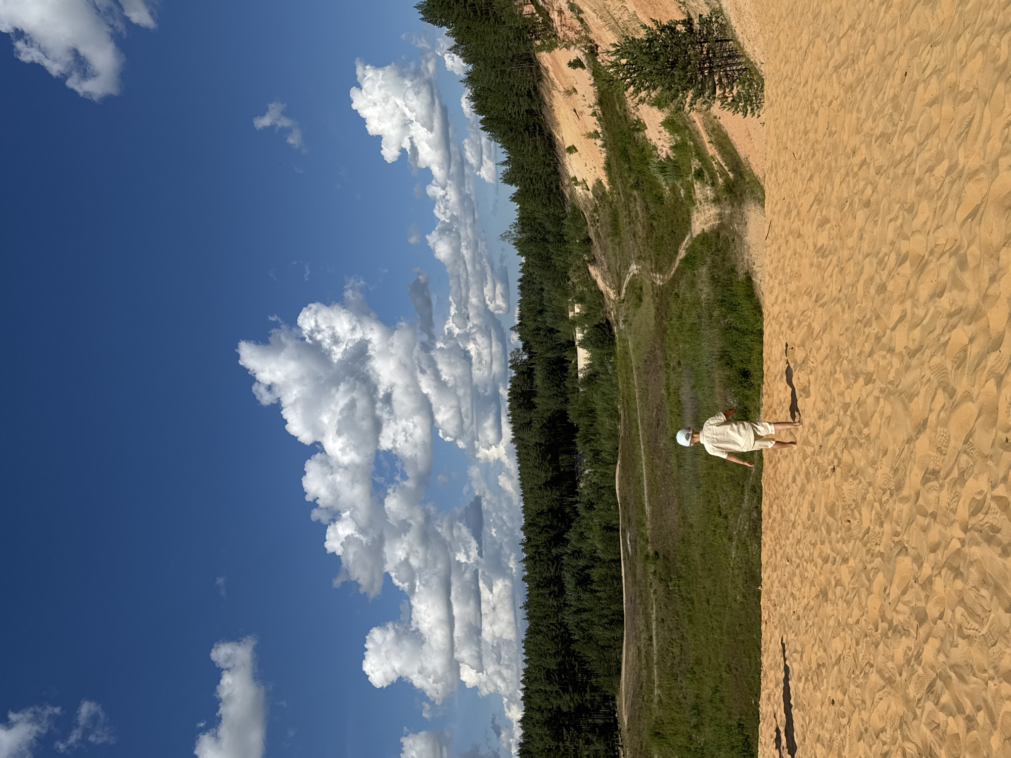 The image shows a wide shot of a sandy landscape with a young boy walking away from the camera. The boy is wearing a light-colored shirt and shorts and a white cap. He is walking on a large sand dune, and in the background, there is a forest and a clear blue sky with white clouds. The sand dune is golden brown and textured with footprints. The forest in the background is dense and green, with a mix of coniferous trees. The sky is a vibrant blue with fluffy white clouds scattered across it. The overall scene is one of a bright, sunny day in a natural outdoor environment. The location appears to be the Piusa Sand Caves area in Piusa, Estonia, known for its large sand dunes and sandy terrain.