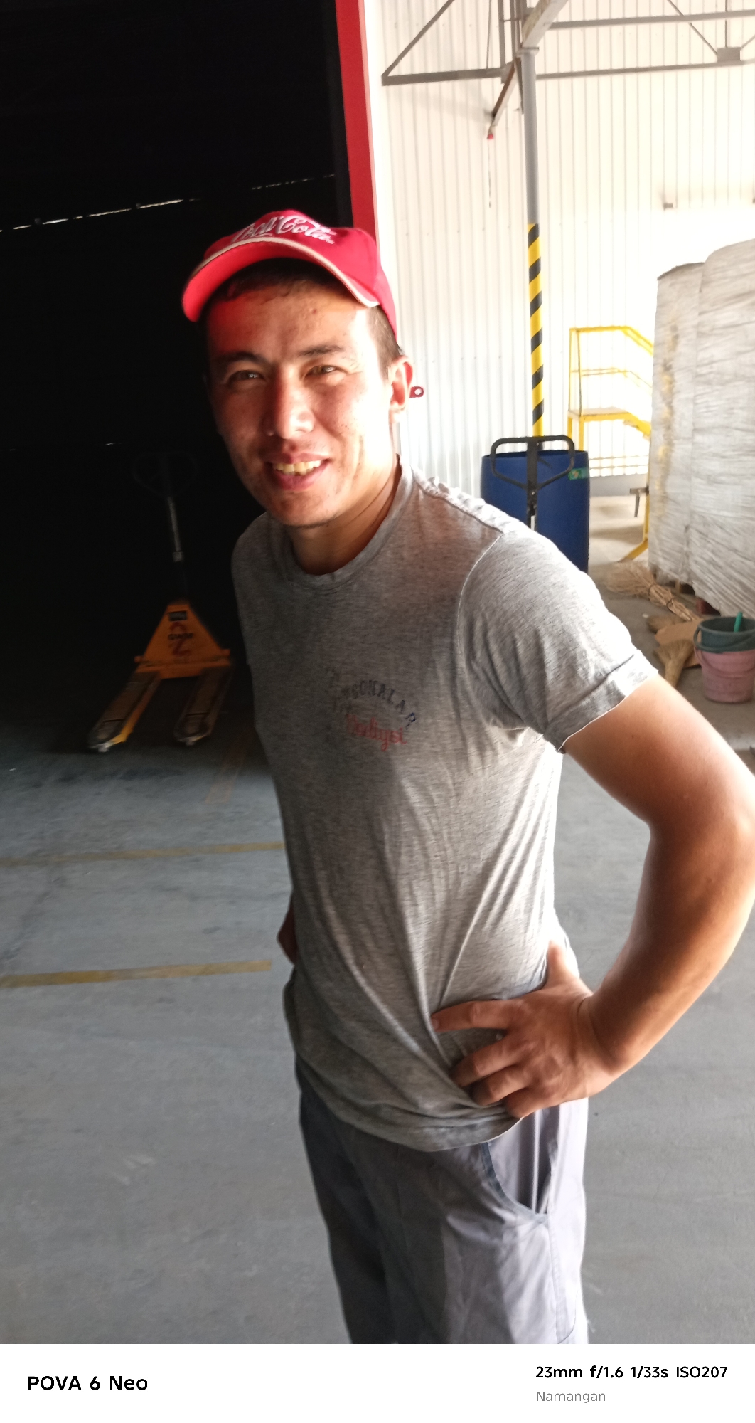A medium shot captures a man wearing a red Coca-Cola baseball cap and a gray t-shirt, posing with his hand on his hip in what appears to be a warehouse or industrial setting. The man is looking directly at the camera and smiling. In the background, a yellow pallet jack is visible in the dimly lit left side of the frame, while the right side shows a large roll of material wrapped in plastic. The floor is concrete with yellow markings. The text overlays indicate the image was taken in Namangan City, Uzbekistan, with the camera settings 23mm f/1.6 1/33s ISO207, and the device POVA 6 Neo.