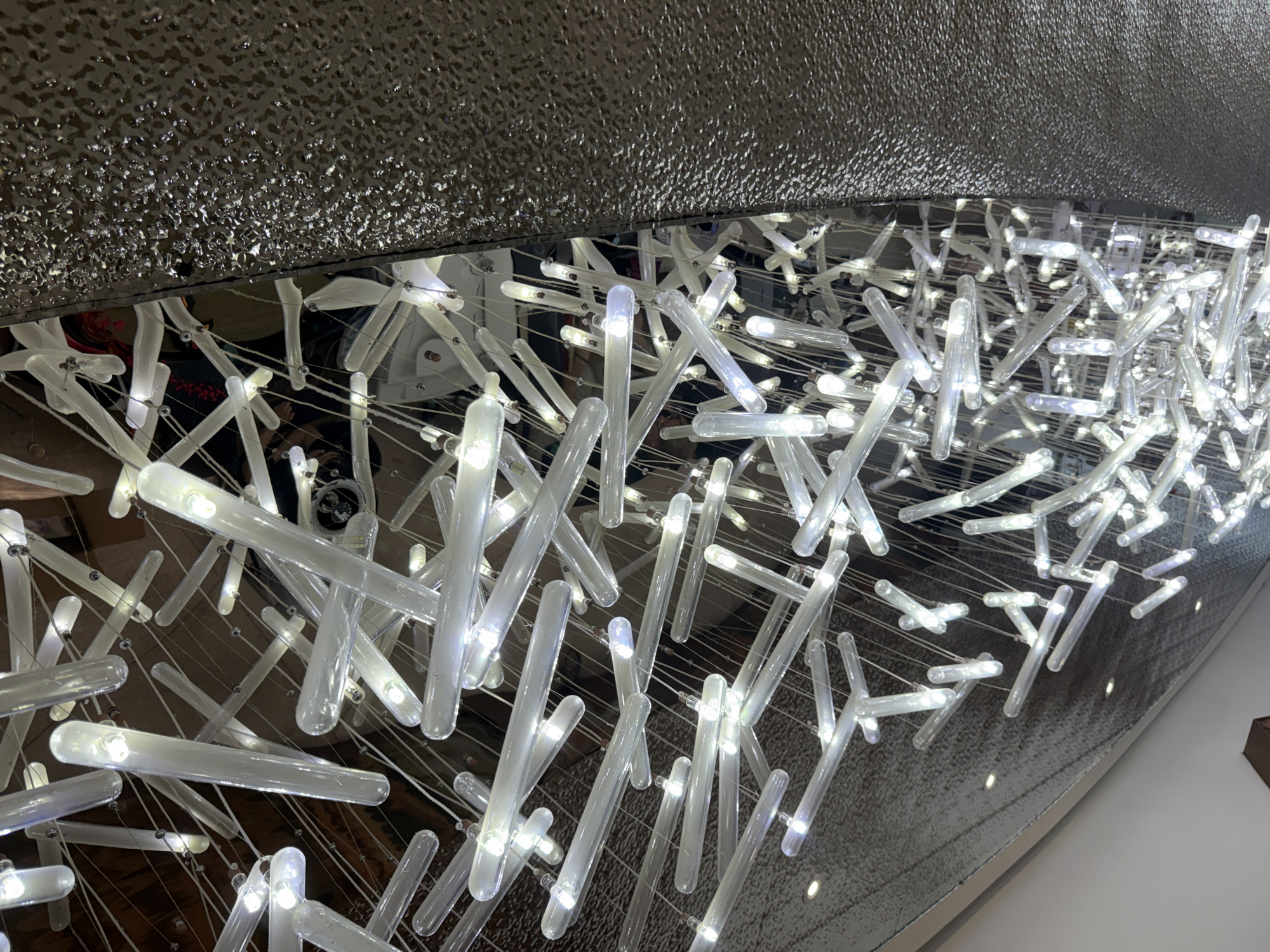 The image shows a close-up view of a modern chandelier made of numerous clear glass tubes containing LED lights. These tubes are suspended by thin wires, creating a complex and layered effect. The lights are illuminated, casting a bright, cool glow. The background is a mix of reflective surfaces, including what appears to be a metallic wall or ceiling with a textured pattern. Some darker areas suggest reflections of the environment above and around the chandelier, possibly including hints of people or other architectural elements. The overall impression is one of contemporary, artistic interior design. The image was captured in Lin'an District, China, though no specific location within the district is identifiable from the visual information provided.