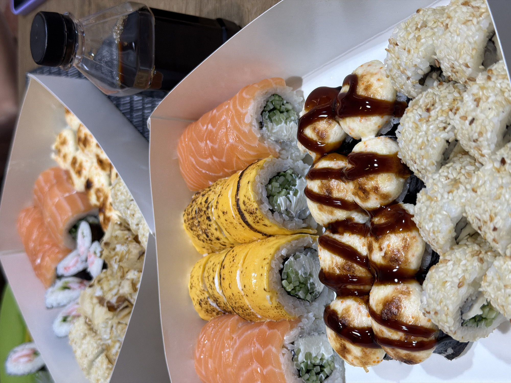 The image displays an overhead view of several sushi containers, a bottle of soy sauce, and a textured dark placemat. The sushi containers, made of white cardboard, are filled with various types of rolls. In the foreground, one container holds sushi rolls with a prominent topping of baked crab or fish salad drizzled with dark sauce, and other rolls are coated in sesame seeds. In the background, another container contains salmon sushi rolls and rolls with a bright yellow outer layer, possibly omelet or cheese, which are also lightly browned. The bottle of soy sauce is positioned to the right, partially obscuring the background sushi container. The overall setting appears to be a table. The provided context does not contain any information about the location in Khmelnytskyi, Ukraine, nor are there any people or discernible actions depicted.