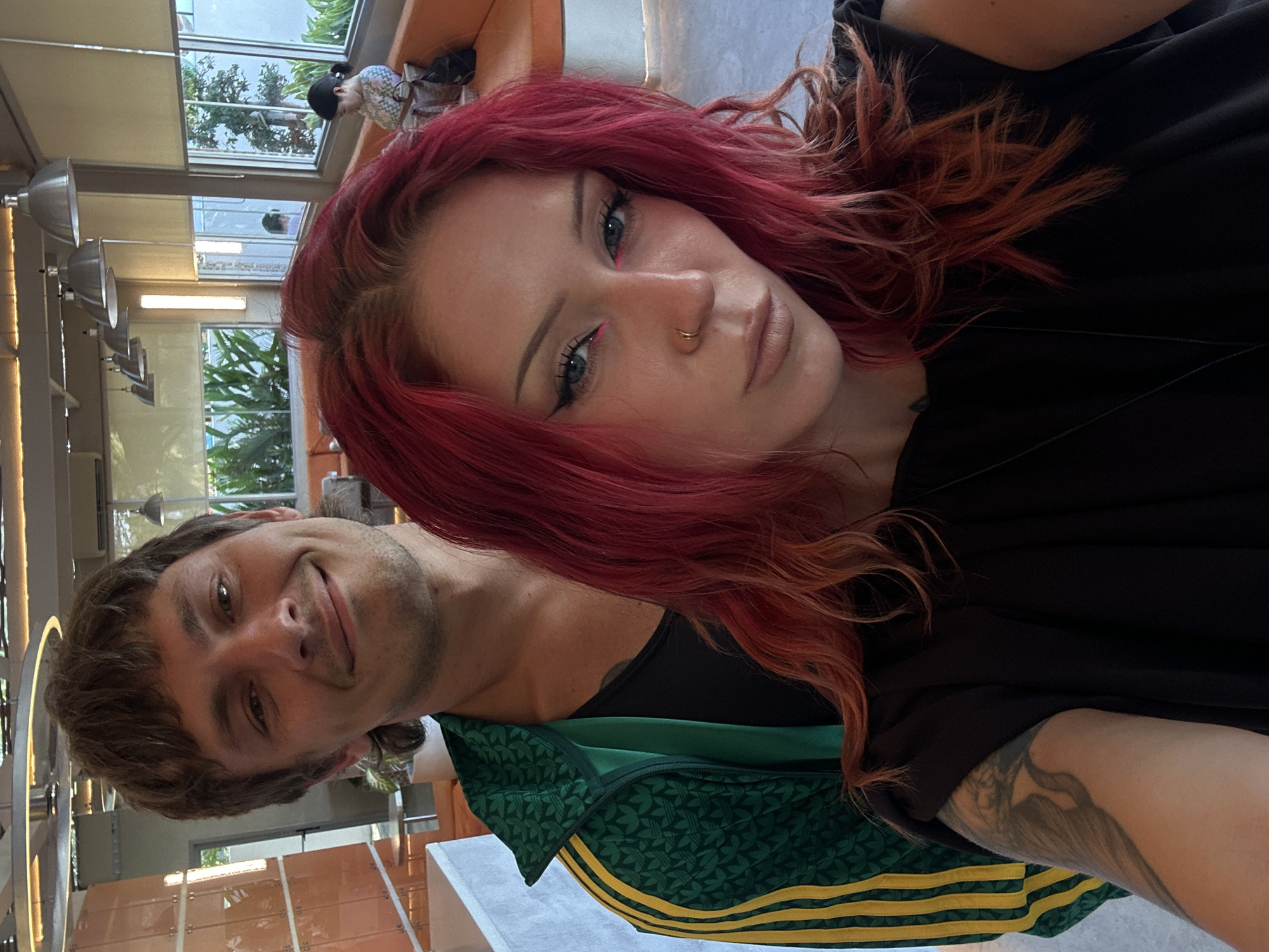 A selfie photograph shows two individuals. The female, positioned in the foreground, has red hair and wears a black top. She has light skin and is wearing pink eyeliner and a nose ring. The male, located slightly behind her and to the left, has brown hair and a slight smile. He is wearing a green and yellow Adidas jacket. The image is taken indoors, with architectural elements and other people visible through a window in the background. The location is likely in North Kuta, Indonesia.