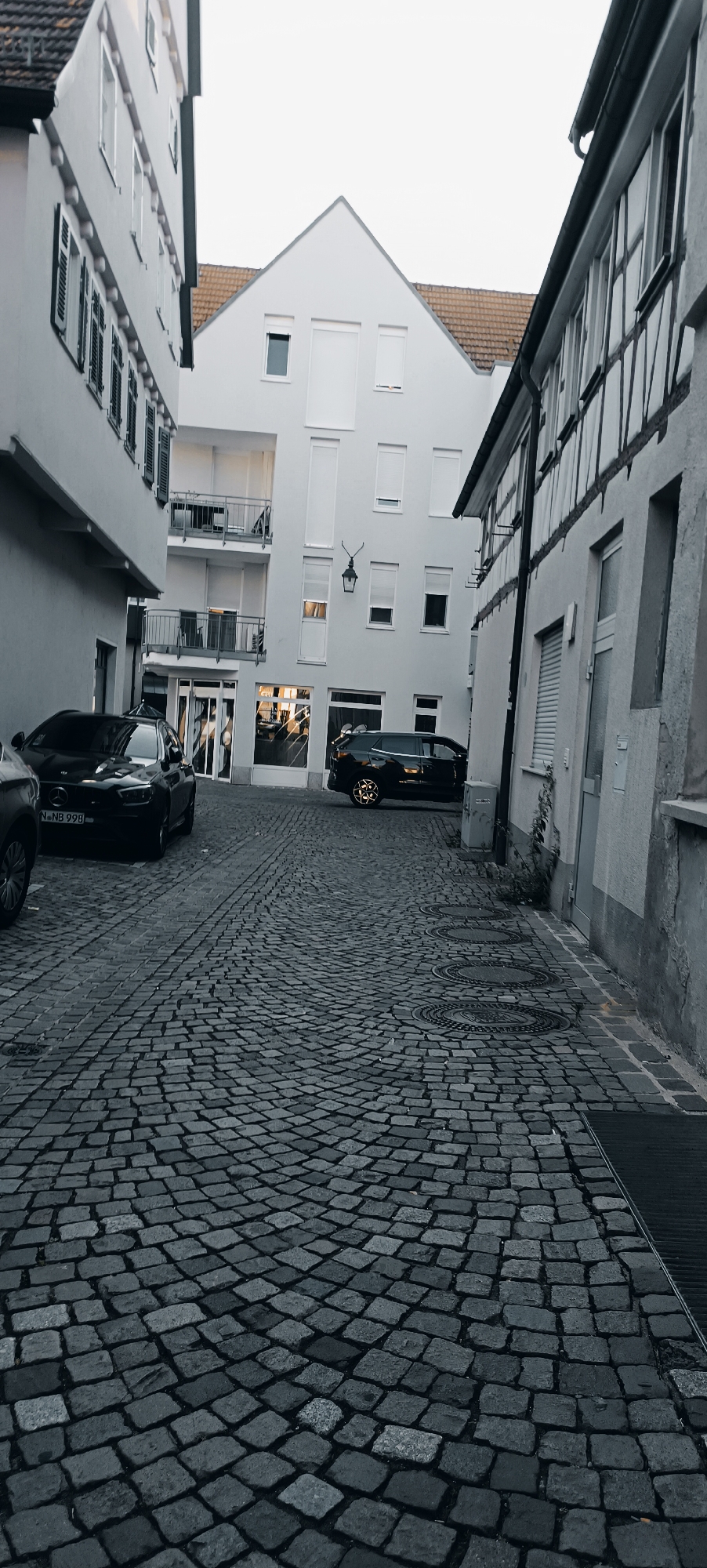 This is a medium-shot, eye-level, grayscale photograph of a narrow cobblestone street in Winnenden, Germany. The street is lined with buildings on both sides. On the left, a light-colored building with shutters is visible. On the right, a building with a half-timbered facade is present. In the background, a white modern building with multiple floors and balconies is situated. Two black cars are parked on the street. One car, a Mercedes-Benz, is parked on the left side of the street, facing towards the viewer. The second car, an SUV, is parked on the right side of the street, facing away from the viewer. The street is paved with cobblestones, and there are several manhole covers visible in the foreground. The sky is overcast. The scene appears to be captured during daylight, likely in the late afternoon or early evening, given the lighting conditions and the absence of strong shadows.