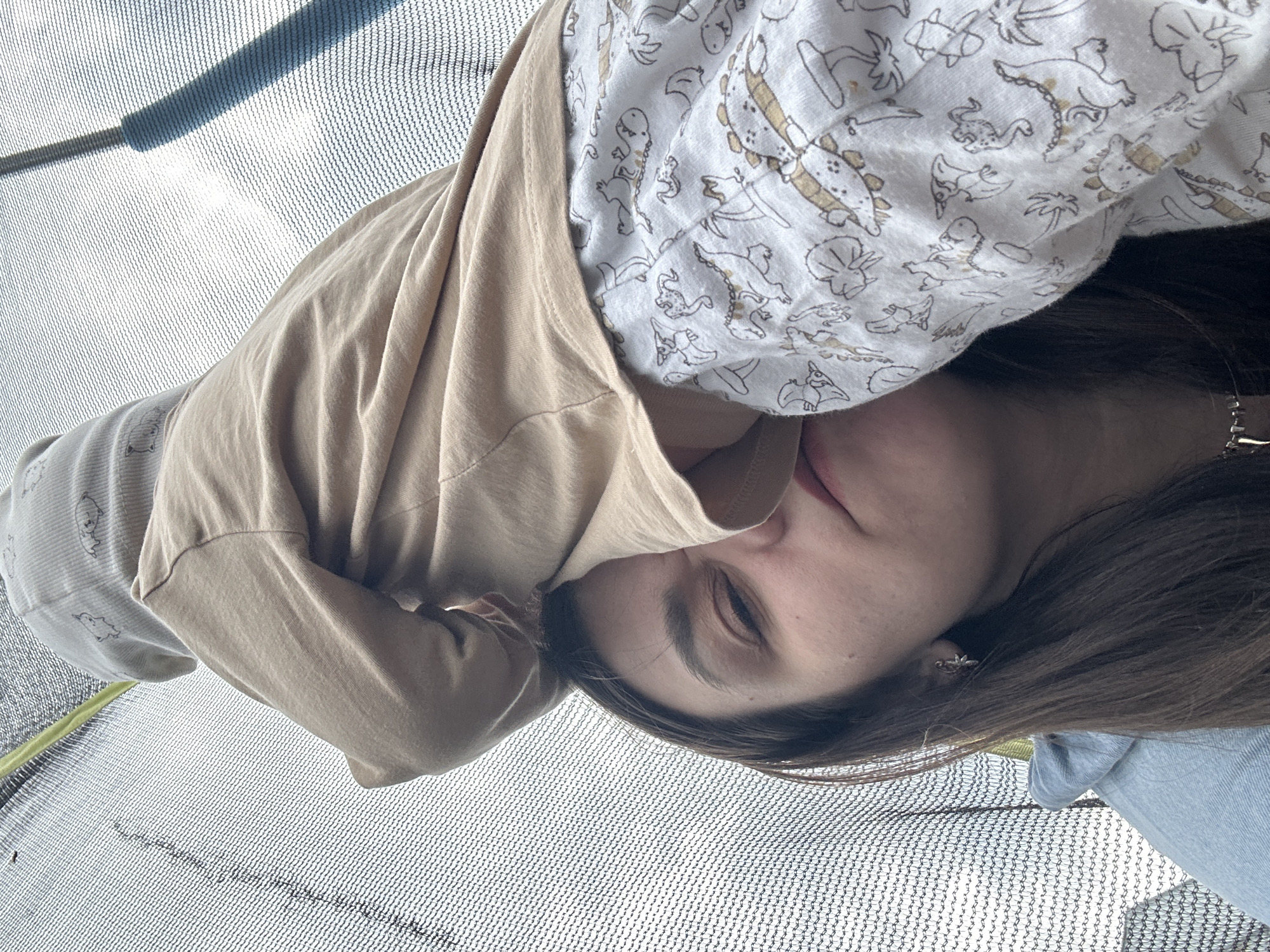 A woman lies on a trampoline. She is wearing a beige sweatshirt over a white shirt patterned with dinosaurs. Her brown hair is loose, and she has a small earring. The trampoline netting surrounds her. The location is unknown.