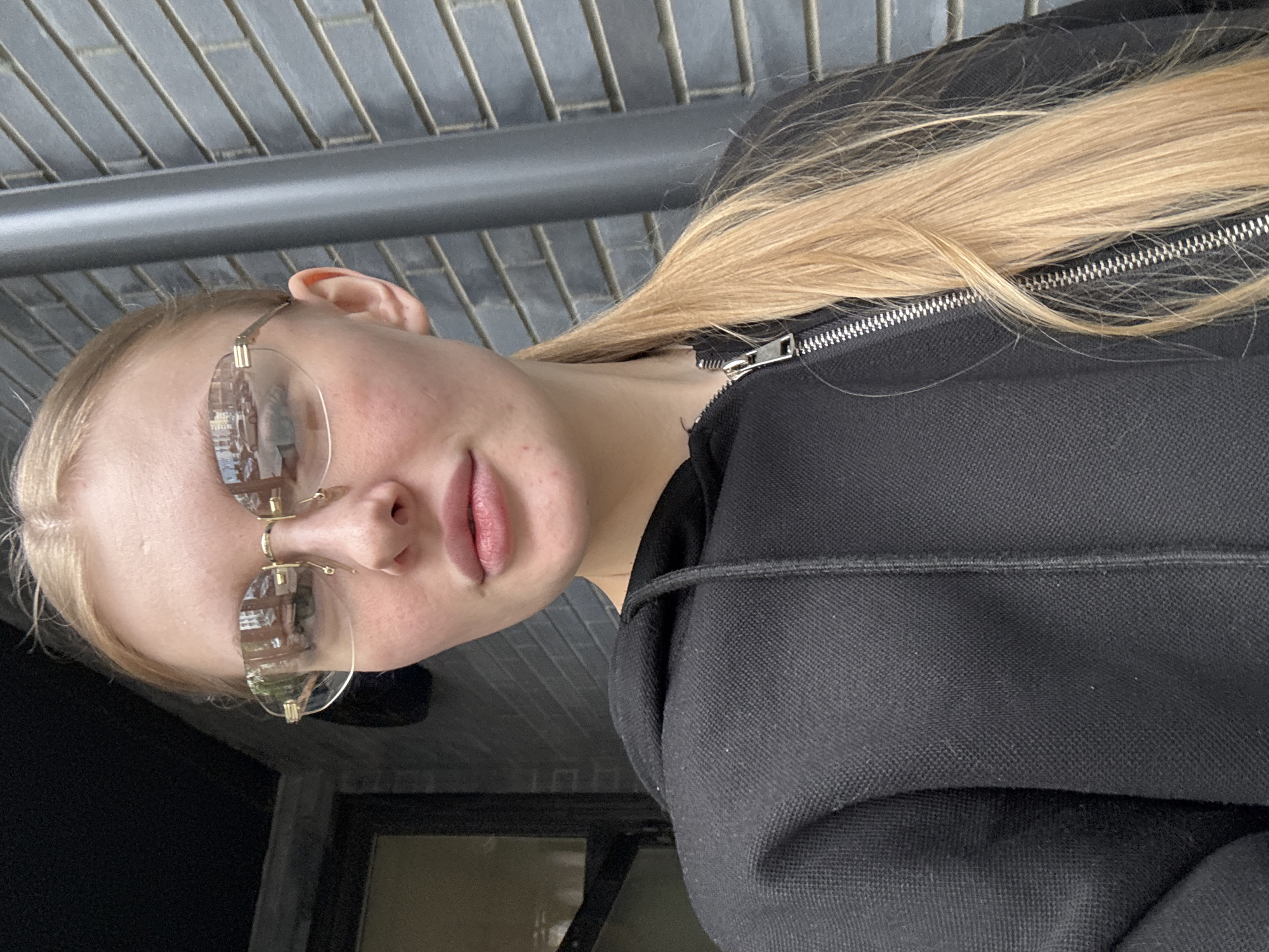 A woman with blonde hair is wearing glasses and a black jacket. The reflection in her glasses suggests she is in Perm, Russia. The background shows a building with windows.