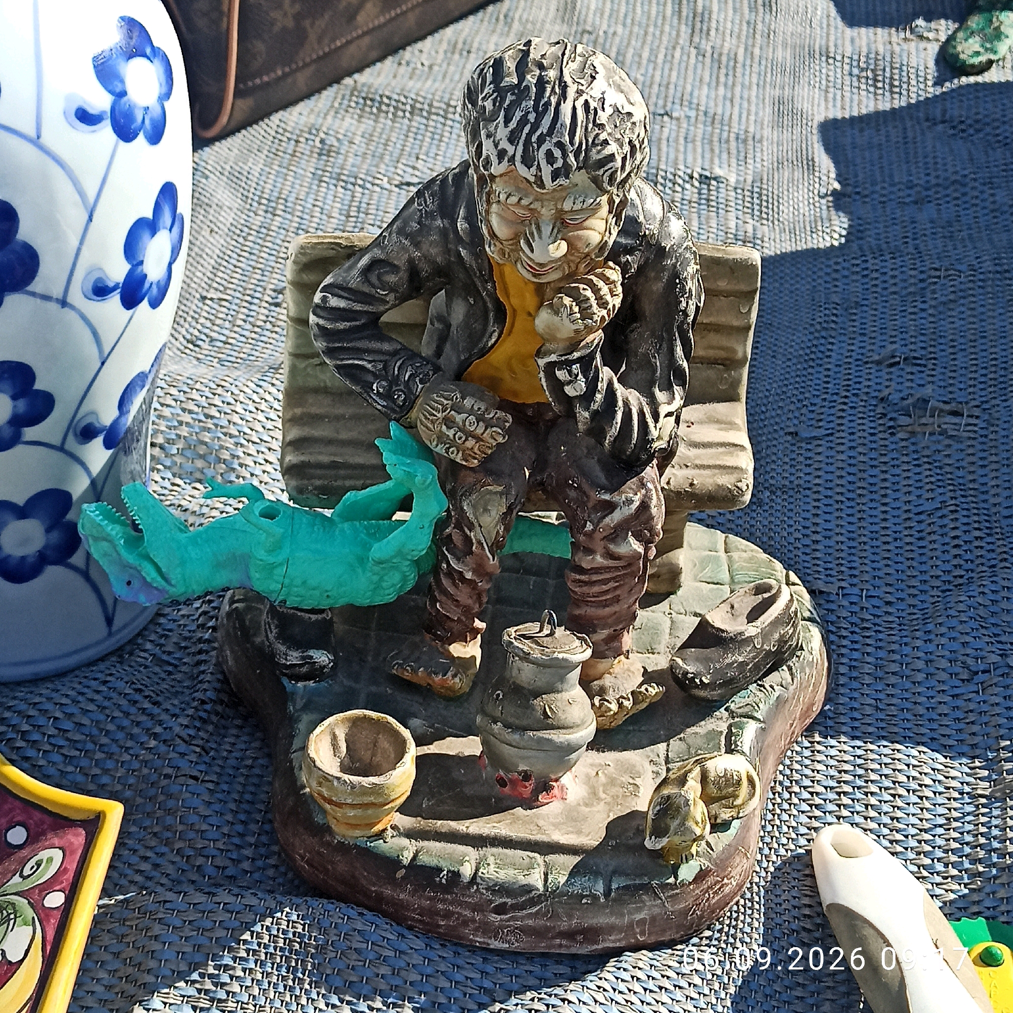 The image depicts a figurine of a disheveled man sitting on a bench, holding his chin in contemplation. Beside him, a turquoise dinosaur-like toy is positioned. The figurine is placed on a textured, blue surface, possibly a mat or fabric. In the background, a portion of a blue and white patterned vase is visible. The scene appears to be outdoors, under natural light, with shadows cast on the surface. The date stamp "06.09.2026 09:17" is visible in the bottom right corner. There is no visible indication of the specific location within the city of Sannicandro di Bari, Italy.
