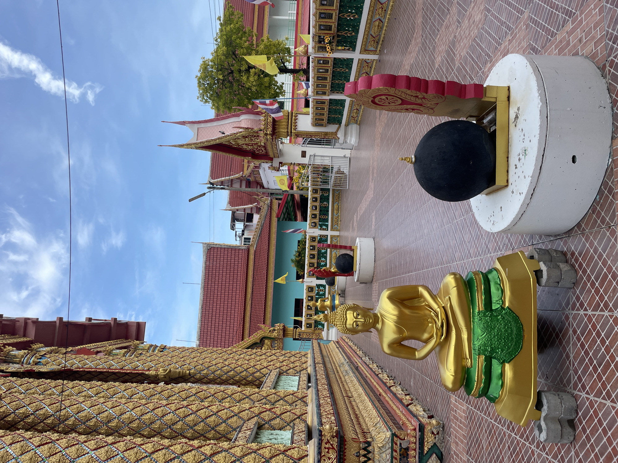 The image shows a Buddhist temple in Pattaya City, Thailand. In the foreground, a golden Buddha statue sits on a green and gold pedestal, which is elevated by gray concrete blocks. To the right of the Buddha statue is a large black sphere on a white pedestal, with a red and gold decorative element behind it. The ground is paved with red-brown tiles arranged in a diamond pattern. In the background, the temple buildings have red roofs and ornate golden decorations on their walls. There are flags of Thailand displayed, as well as yellow flags. The sky is blue with scattered clouds. The scene appears to be outdoors, in a courtyard or walkway of the temple.