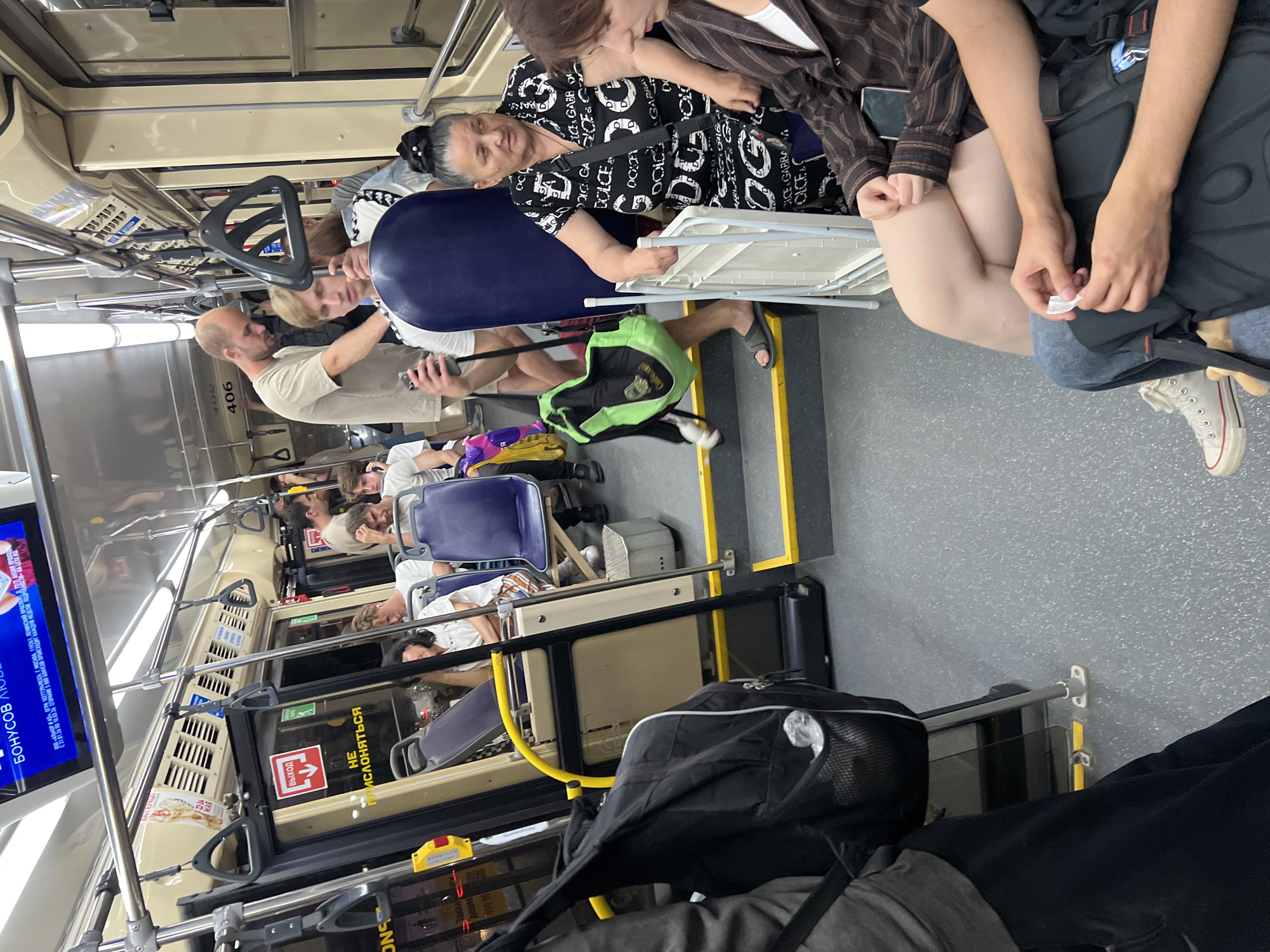 The image depicts the interior of a public bus, likely in Krasnodar, Russia, as suggested by the presence of a sign with Cyrillic script indicating "ВЫХОД" (Exit). The bus is moderately crowded, with passengers seated and standing. In the foreground, a black backpack with a mesh pocket is visible, resting on the floor. To the right, a woman with dark hair pulled back in a bun is seated, wearing a black and white patterned dress. She is holding a folded white object, possibly a ticket or piece of paper. Next to her, a person with light brown hair is seated, wearing a striped jacket.
In the middle ground, several passengers are visible. A man in a light-colored t-shirt is standing, holding a phone. A young man with blonde hair is seated in a blue seat, looking down. Another man in a light-colored t-shirt is standing and looking forward. A green backpack is visible on the steps. Further back, a man with a shaved head is standing, and another person is seated.
The bus interior features typical public transport elements such as overhead handrails, blue and beige seats, and yellow safety markings on the steps and floor. A digital display board is mounted above the passengers, showing a blue screen with text, partially obscured. The overall environment appears to be daytime, with artificial lighting from the bus interior visible. The atmosphere seems ordinary for a bus commute.
