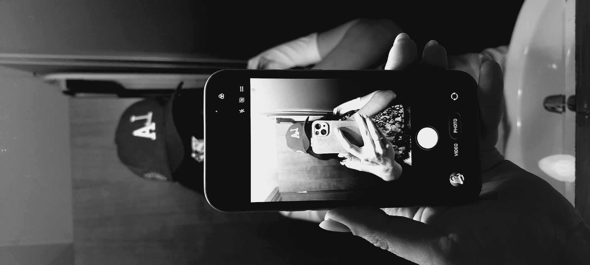 This black and white, vertically oriented photograph captures a selfie taken in what appears to be a bathroom. The primary subject is a person, whose face is obscured by the phone they are holding. They are wearing a baseball cap with a white "LA" logo on the front. The person is holding a smartphone in their right hand, angled to take a selfie. The phone's screen displays the camera interface, showing a reflection of the person, the cap, and a floral-patterned garment. Visible on the phone screen are camera mode options like "VIDEO" and "PHOTO," zoom levels (0.5x, 1x), and a circular shutter button.
The setting seems to be a bathroom, suggested by the presence of a white sink with a faucet visible in the background on the right side of the image. The lighting is somewhat dim, casting shadows and creating a dramatic effect typical of indoor settings with limited light. The overall mood is casual and perhaps a bit playful, indicated by the selfie pose. The image is clean and sharp, focusing on the phone and the immediate surroundings. There is no information about the time of day or weather. The location context is provided as Sofia, Bulgaria.