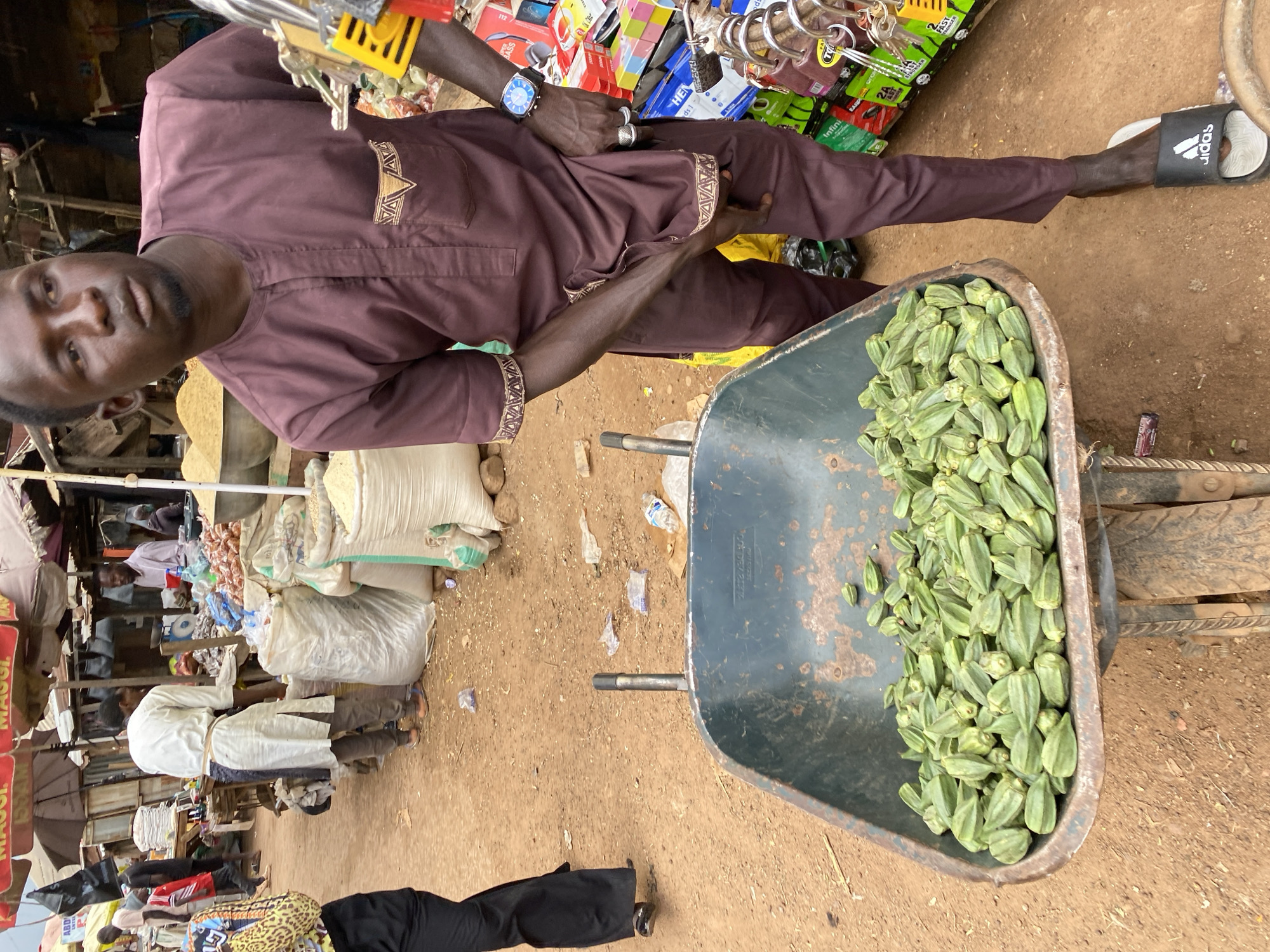 A man, possibly a vendor, stands in a bustling outdoor market in Bukuru, Nigeria, with a wheelbarrow filled with okra in the foreground. He is wearing a maroon traditional outfit and a wristwatch. The wheelbarrow, an old and weathered industrial tool, is positioned centrally in the frame, its contents spilling slightly over the sides. The market environment is visible behind the man, with stalls displaying various goods, sacks of grains, and other vendors in the background. The ground is sandy and uneven, typical of an outdoor market. The overall scene depicts a snapshot of daily commerce in a Nigerian market.