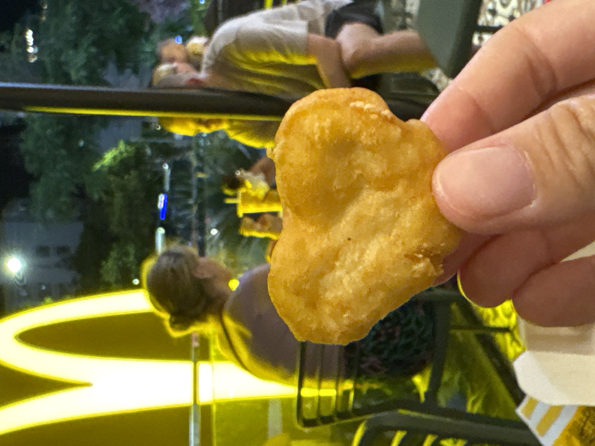 An outdoor dining area at a McDonald's restaurant in Nha Trang, Vietnam, is depicted at night. In the foreground, a hand holds a chicken nugget. In the background, people are seated at tables. A large, illuminated McDonald's logo is visible on the left side of the image. The scene appears to be a typical fast-food restaurant setting.