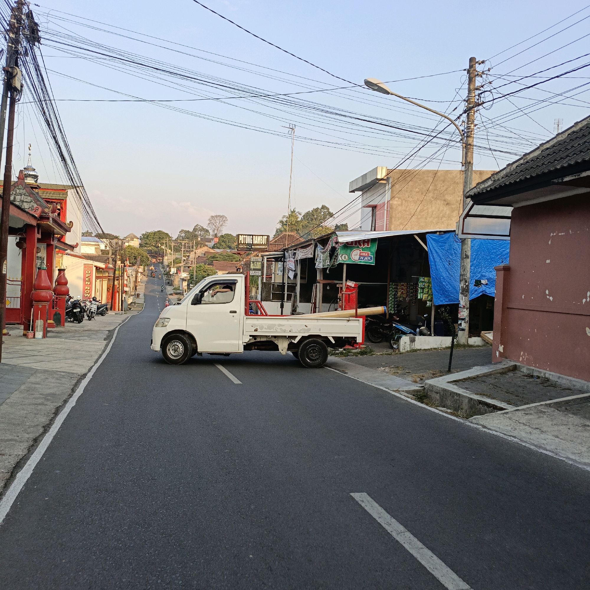 The image depicts a street scene in Salatiga, Indonesia, likely during late afternoon or early evening due to the soft, warm light. A white pickup truck is driving on the right side of the asphalt road. The truck has a red frame around its cargo bed and is carrying a long, light-colored object, possibly piping or a rolled material. The truck is partially obscuring a row of shops and businesses on the right side of the road. One shop has a blue tarp covering its front, and another appears to be a barber shop with a sign reading "POTONG RAMBUT" (haircut).
On the left side of the road, there is a building with red accents and an ornate roof, resembling a temple or traditional religious structure. Several motorcycles are parked on the sidewalk in front of this building. Further down the street, there are more shops, buildings, and a gentle incline. Utility poles with a complex network of wires are prominent throughout the scene, typical of urban infrastructure. The sky is clear and pale blue, suggesting fair weather. The overall impression is a snapshot of everyday life on a commercial street in Salatiga.