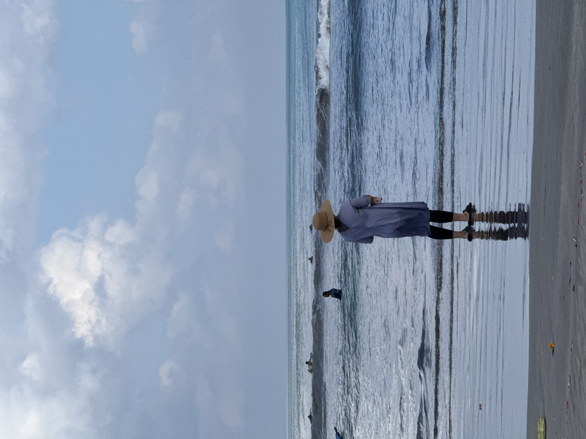 A person wearing a light blue dress and a wide-brimmed hat stands on the wet sand of a beach, facing the ocean. The person's reflection is visible in the shallow water covering the sand. In the background, several surfers are in the water, some on surfboards and others paddling. The sky is cloudy, with patches of blue showing through. This scene is consistent with the typical scenery at Kuta Beach in Bali, Indonesia, known for its surfing conditions and popularity with tourists.