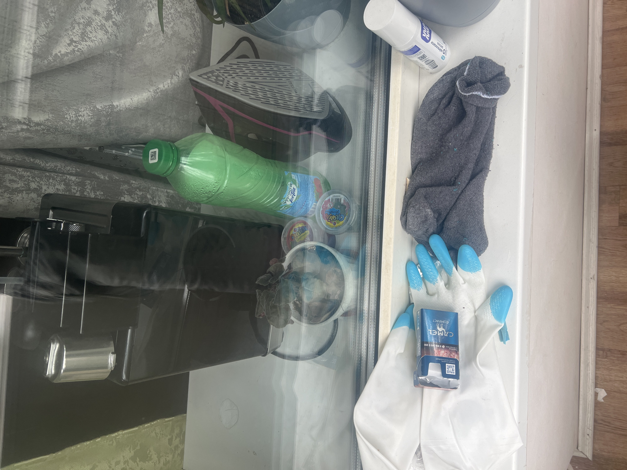 The image shows a windowsill in Lobnya, Russia. On the windowsill are several items, including a white and blue gardening glove, a pack of Camel Compact cigarettes, a gray sock, a white spray can, and a green bottle of tea. Behind these items, reflected in the glass, are a black coffee machine, a black iron, and a potted plant. The overall impression is one of a cluttered windowsill with a mix of everyday items and cleaning supplies.