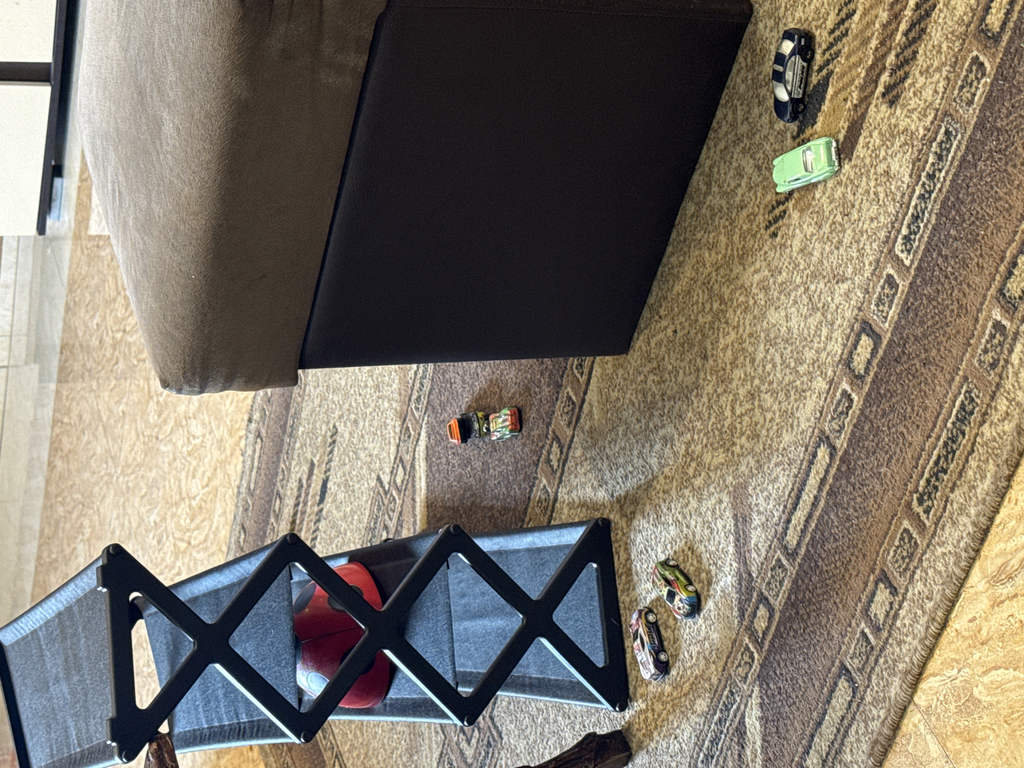 The image shows an indoor scene on a brown patterned carpet. A section of a brown and black upholstered piece of furniture is visible in the upper left. Several toy cars are scattered on the carpet. A black expandable barrier containing a red ball is in the lower part of the image. There are no people visible. There is no information given in the photo that can be used to determine a specific location in Yerevan, Armenia.