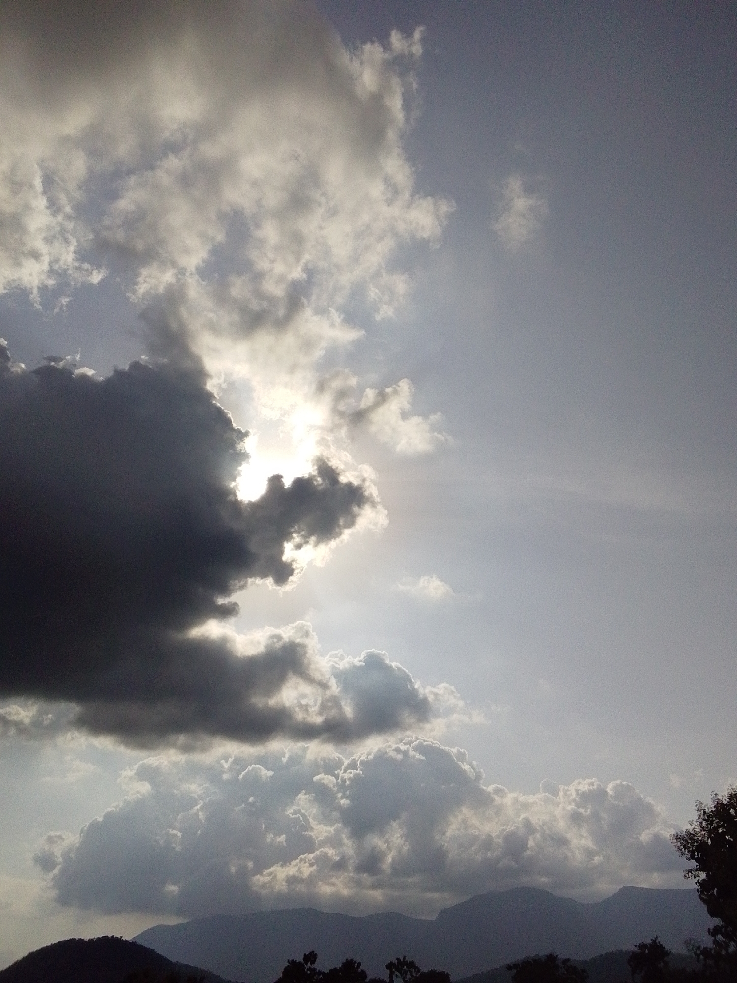This is a vertical, medium shot of a dramatic sky over Serti, Nigeria, likely taken in the late afternoon or early evening as indicated by the low angle of the sun. The sky is filled with a mixture of dark, heavy storm clouds and lighter, more diffuse clouds. The sun is partially obscured by a large, dark cloud formation in the upper left quadrant of the image, casting a bright, ethereal glow through the edges of the clouds.
In the lower part of the image, silhouetted against the sky, are the dark shapes of rolling hills and mountains. The foreground also shows the dark silhouettes of trees and foliage, adding depth to the scene. The overall mood is one of atmospheric intensity, with the contrast between the dark clouds and the bright sunlight creating a sense of awe and perhaps foreboding. The weather appears to be unsettled, with the potential for rain indicated by the heavy cloud cover. There is no visible text in the image. The scene is a natural landscape, devoid of human presence or man-made objects.