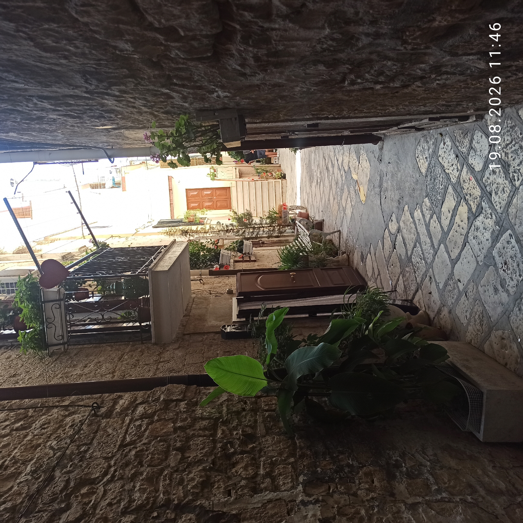 The image depicts a narrow, cobblestone alleyway in the historic center of Ruvo di Puglia, Italy. The walls of the alley are constructed from rough-hewn stone. On the left, a large green plant in a planter is visible. Further down the alley, a white metal bench sits against a building, flanked by potted plants. On the right, the stone wall continues, with a small plant growing from a wooden planter attached to the wall. In the background, a staircase leads up to a wooden door. A date and time stamp, "19.08.2026 11:46", is visible in the lower right corner. The scene appears to be a quiet, residential street.