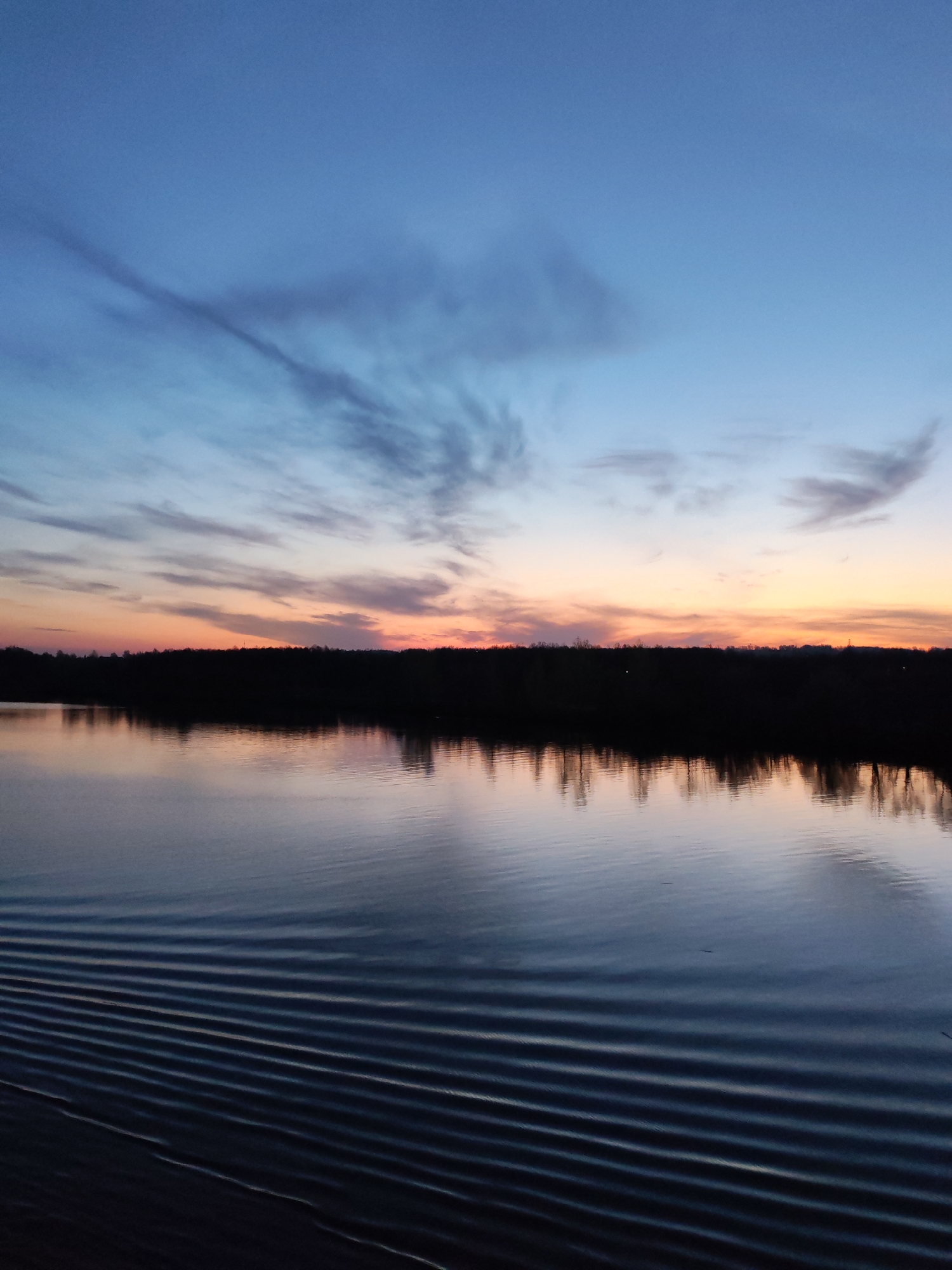 The image depicts a tranquil scene at dusk. A body of water, possibly a lake or river, occupies the foreground. The water's surface is rippled, with noticeable parallel wave patterns extending from the bottom left corner towards the center. These waves suggest the recent passage of a boat or other watercraft.
Above the water, a dark, silhouetted treeline stretches across the middle ground, indicating the presence of land or an island in the distance. The sky above transitions from a deep blue at the top to a warm orange and pink near the horizon. Wispy clouds are scattered across the sky, reflecting the colors of the setting sun. The location is Unknown, Unknown.