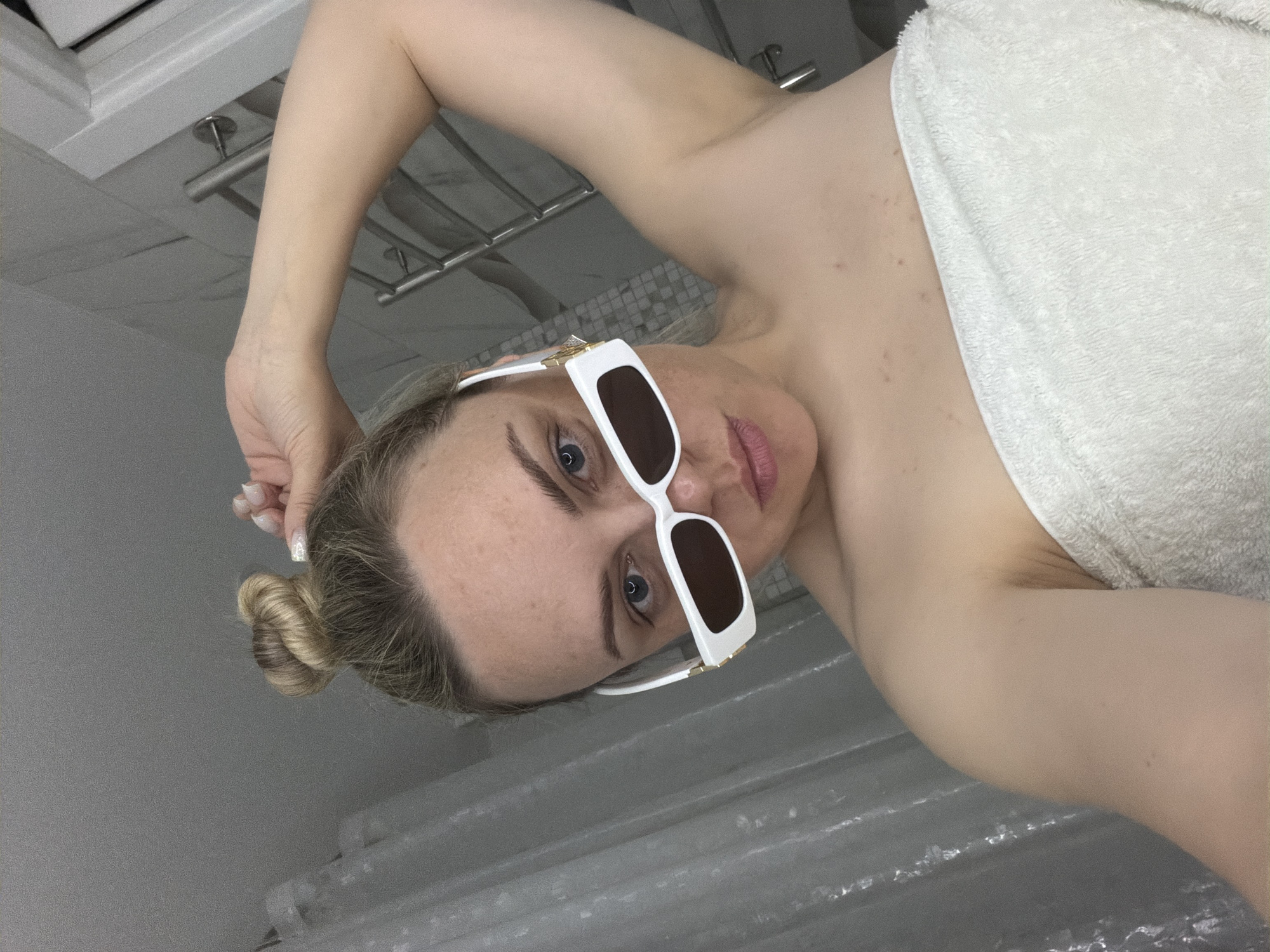 A woman is depicted indoors, presumably in a bathroom based on the presence of a shower curtain, towel rack, and wall tiles. She is partially covered with a white towel and is wearing large white sunglasses. Her hair is styled in a bun. The scene is not located in Votkinsk, Russia.