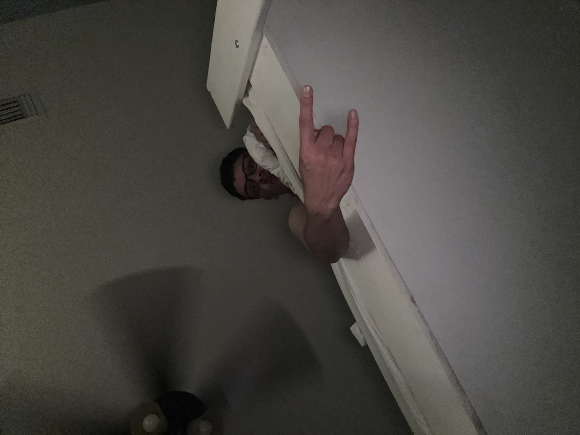 A low-angle shot depicts a person with dark hair and glasses peering down from above. The person is making a hand gesture resembling "rock on" or "devil horns." Part of a white structure, possibly a door frame or wall, is visible, obscuring the person's body. The surrounding environment is dimly lit with gray walls and a floor partially visible. Specific geographical location within Panama City Beach, United States of America, cannot be determined from the available information.