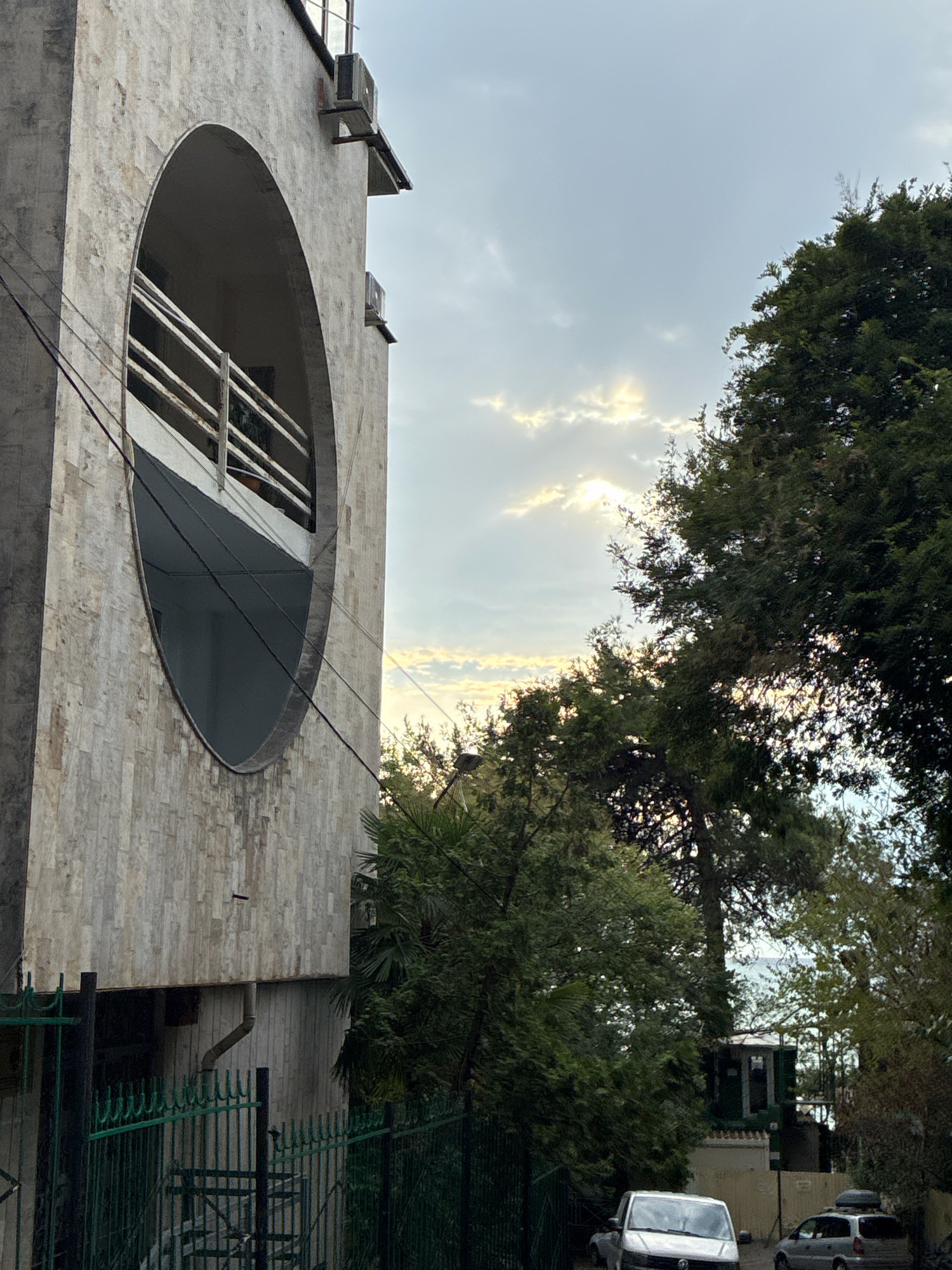 In Sochi, Russia, a low-angle exterior shot captures a building with a prominent circular opening in its facade. The building is clad in light-colored textured tiles. Two air conditioning units are mounted externally on the building's wall. Thick electrical wires traverse the scene in front of the building. To the right, lush green trees dominate the foreground, with sunlight filtering through their branches and illuminating the cloudy sky. In the background, two vehicles are parked on a street, and a glimpse of the sea can be seen through the foliage. A dark green metal fence runs along the bottom left of the image.
