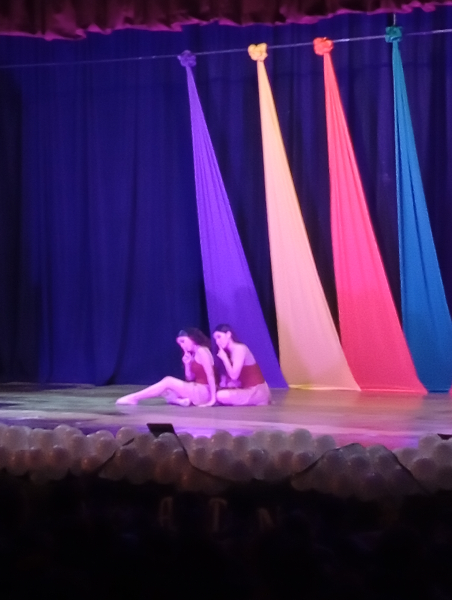 Two female dancers are seated on a stage in Juan Griego, Venezuela. The dancers are wearing matching attire. Behind the dancers, there are draped colorful fabrics. There is an audience present in front of the stage. The edge of the stage is decorated with white objects that may be balloons or some other spherical decor. The stage is lit with theatrical lighting. The backdrop consists of blue fabric.