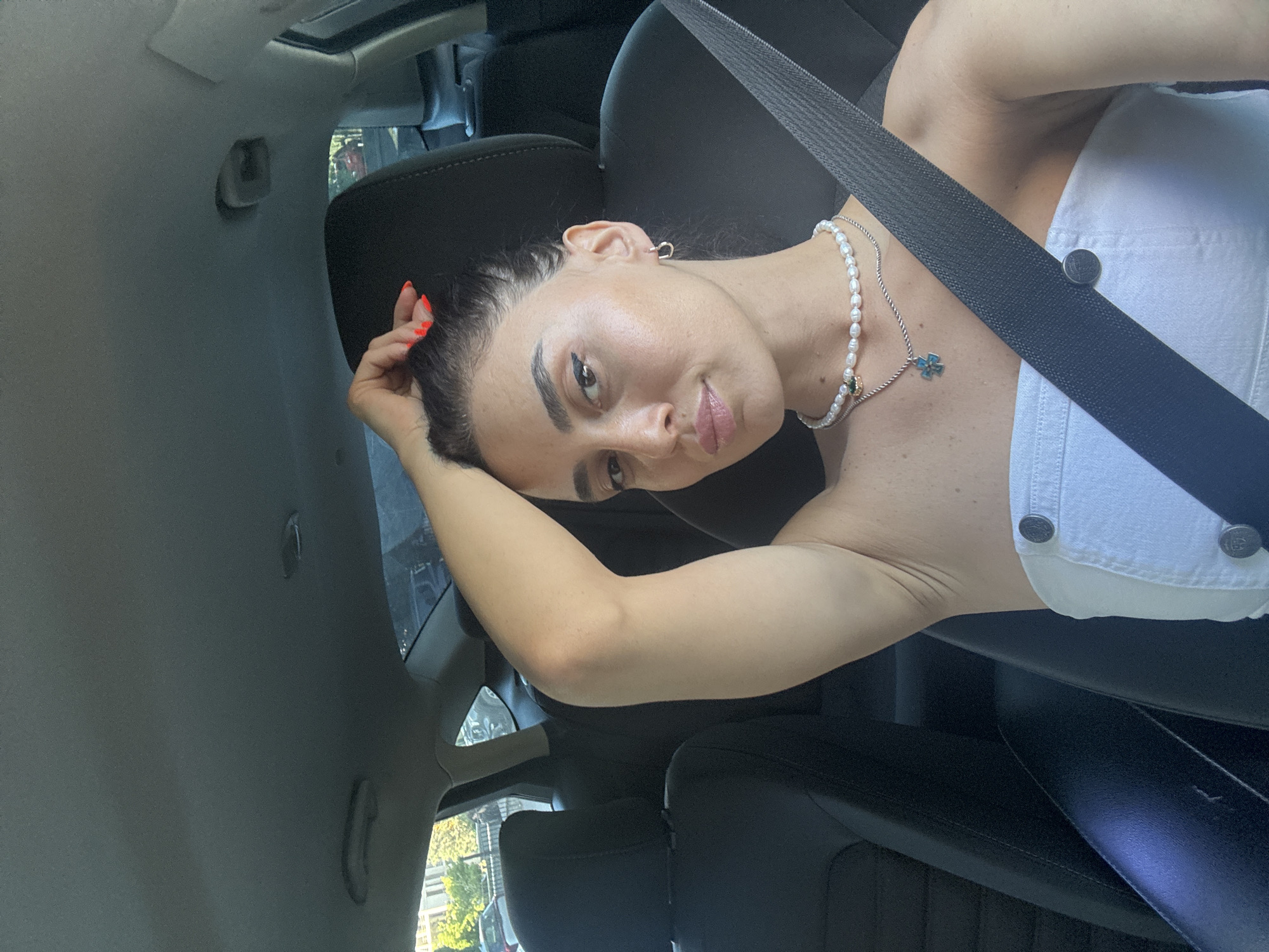 The image shows a woman sitting in the driver's seat of a car. She is wearing a white strapless top and a pearl necklace. Her arm is raised, and she is touching her hair. The car interior is visible, including black leather seats and a seatbelt. The background suggests an outdoor environment with trees and buildings, possibly in a city.