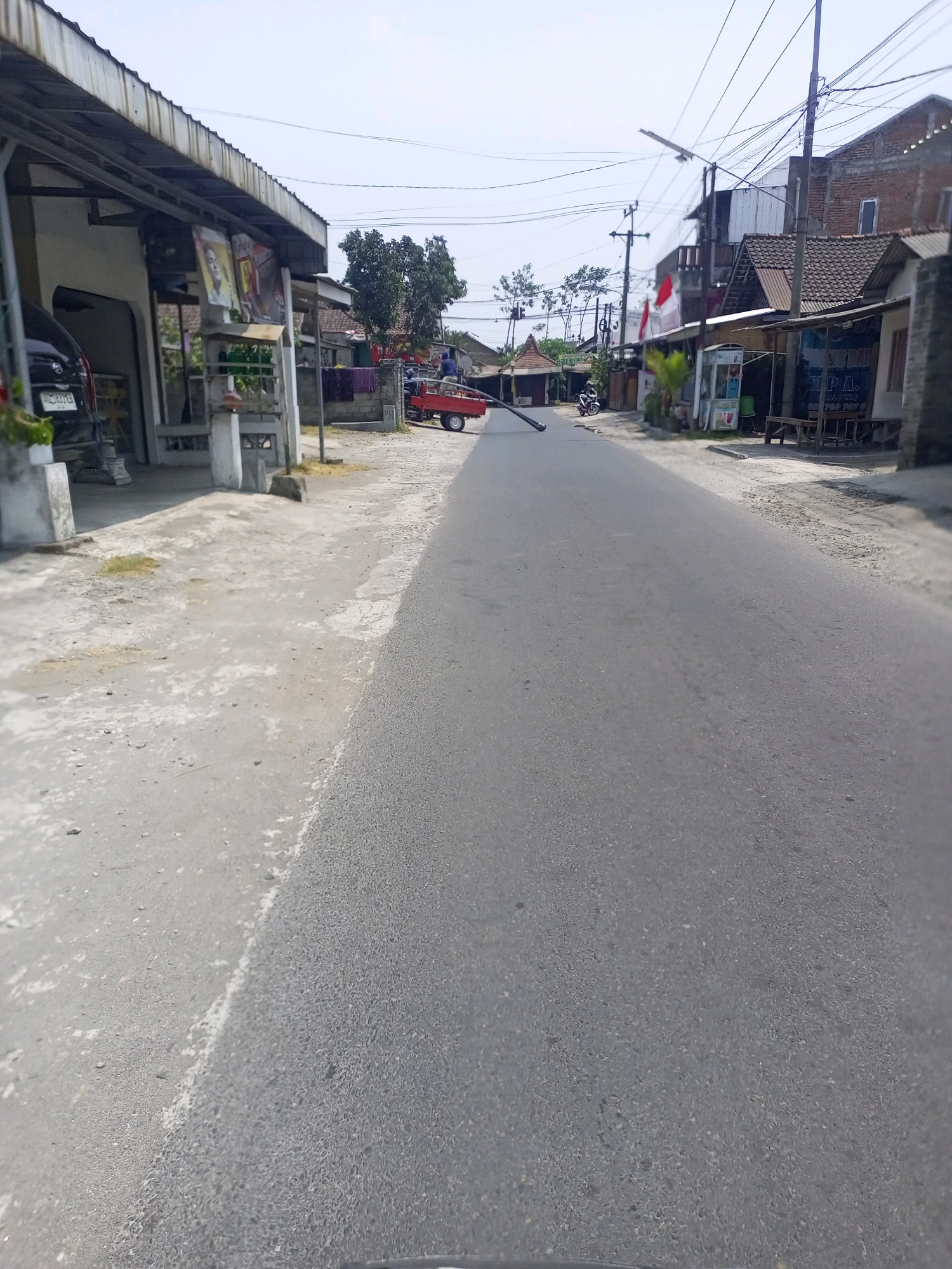 This is a street scene in Ngasem, Indonesia, with a narrow asphalt road running through a village. On the left side of the road, there is a building with a metal awning, a parked black car, and what appears to be a small store or workshop. Further down the left side, there are more buildings and a red truck. On the right side of the road, there are several small shops with tiled roofs and outdoor seating areas. A white and blue vending machine is visible. Overhead, power lines crisscross the sky. The ground is a mix of asphalt and concrete, with some patches of dry grass and dirt. The sky is clear and blue. There are no people clearly visible in the scene.