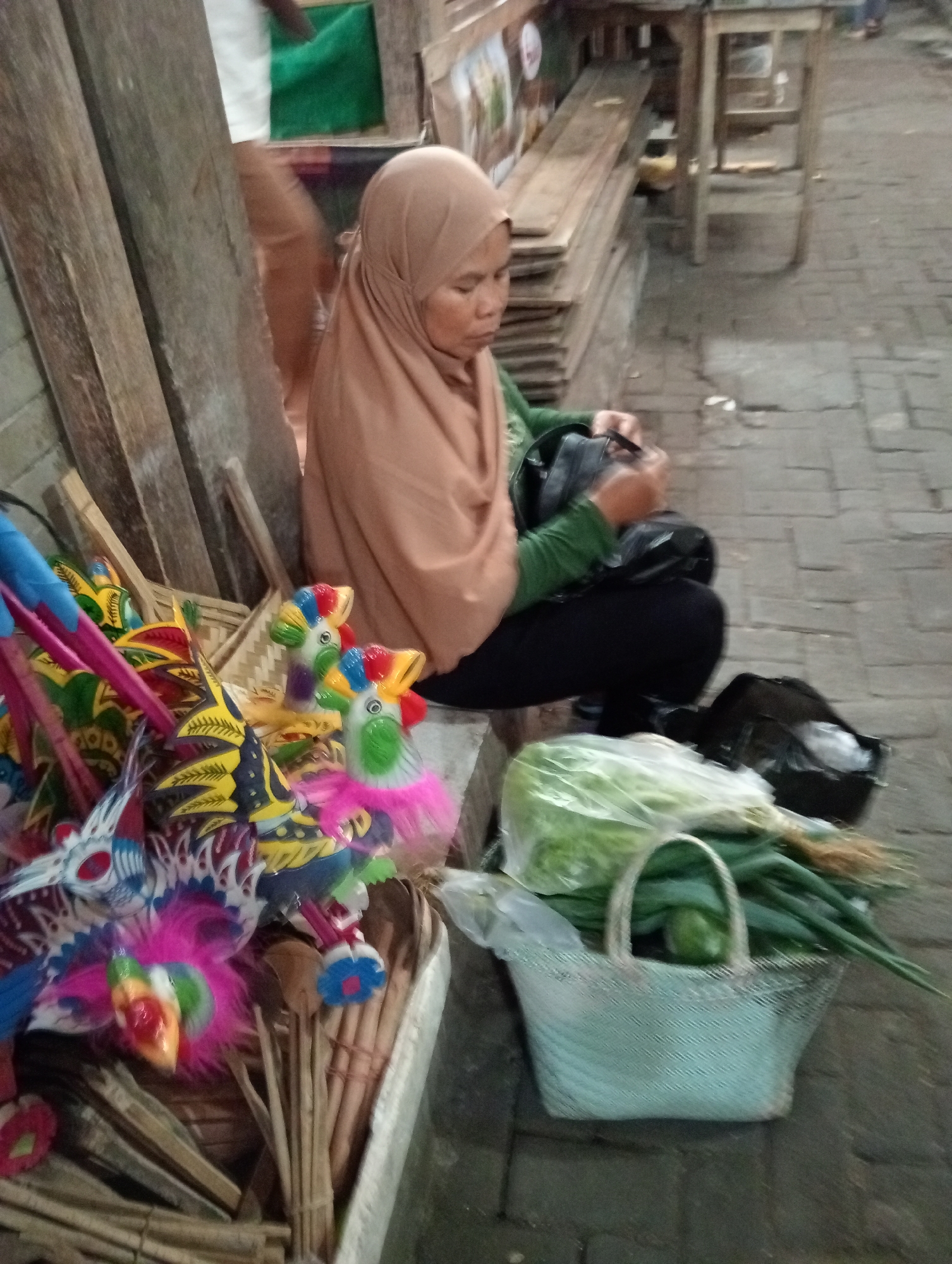A woman wearing a light brown hijab and a green shirt is seated on the ground in Grabag, Indonesia, surrounded by various items. In the foreground, to her left, are several colorful, handcrafted toys resembling birds and dragons, made of wood, paper, and feathers. These toys are arranged in a woven basket. To her right, a light blue woven basket holds fresh green vegetables, possibly scallions and lettuce, wrapped in plastic. Behind the woman, stacked wooden planks lean against a wall. Further in the background, a rustic wooden stall or table is visible, and a person's legs are blurred in motion. The ground appears to be paved with cobblestones. The woman is looking down and appears to be occupied with something in her hands, possibly organizing her goods. The overall scene suggests a market setting, with the woman likely a vendor selling her wares.
